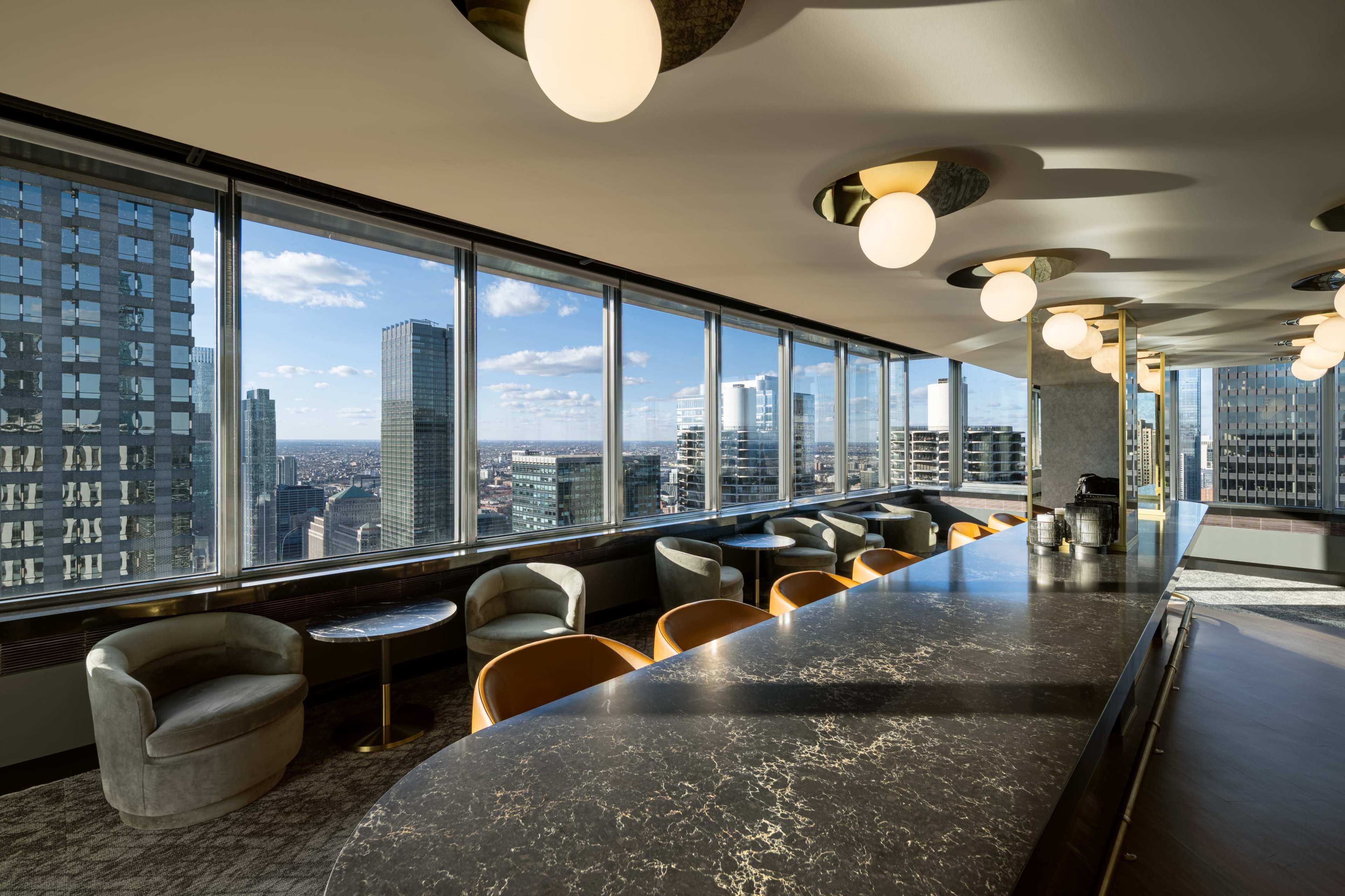 The image shows a modern bar area with a marble countertop and seating, featuring large windows that offer a panoramic view of a city skyline.
