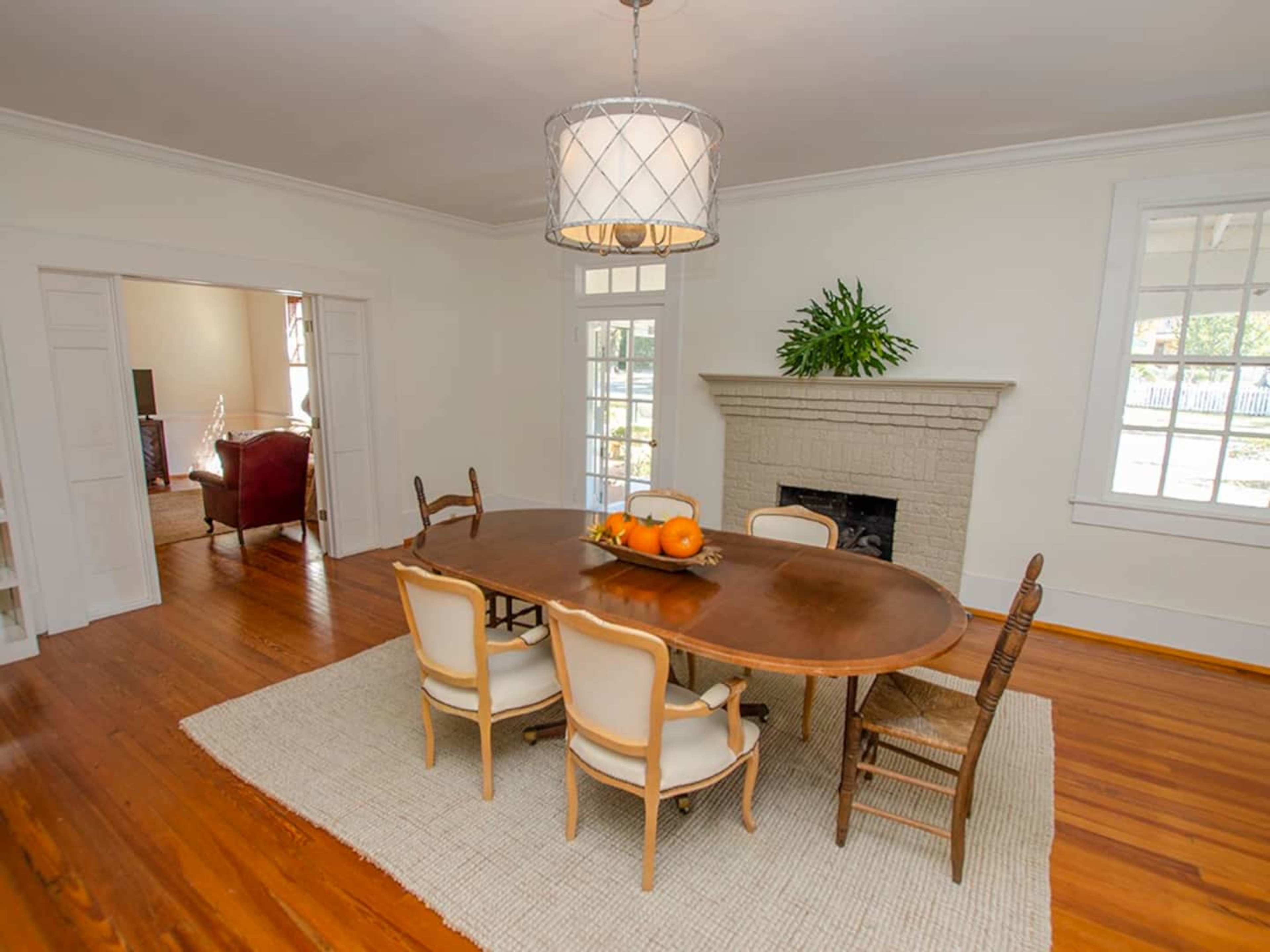 A dining room with a large wooden table surrounded by six chairs, a light fixture hanging above, and a fireplace along one wall.