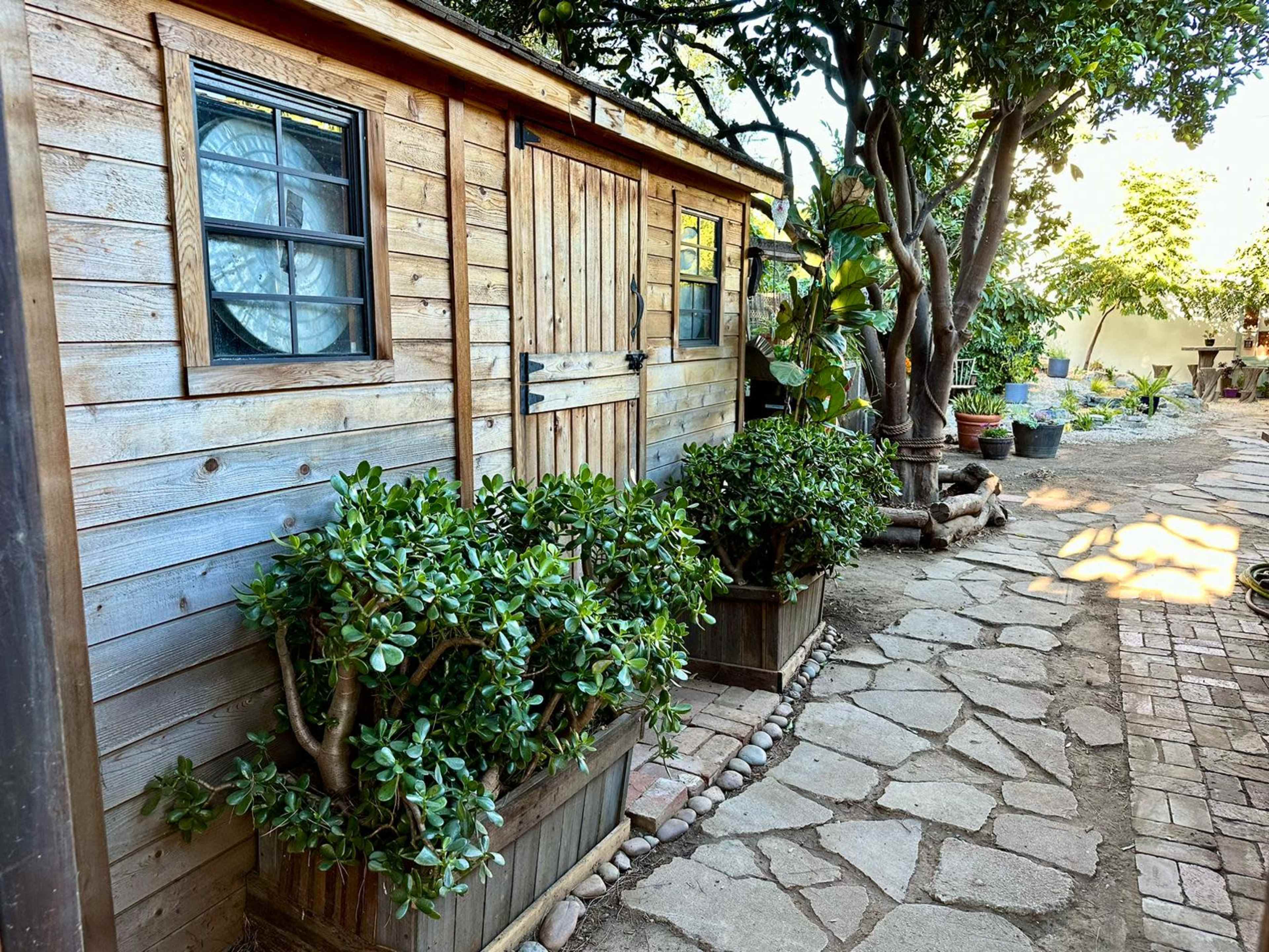 A wooden garden shed is flanked by potted plants and a stone pathway under a canopy of trees.