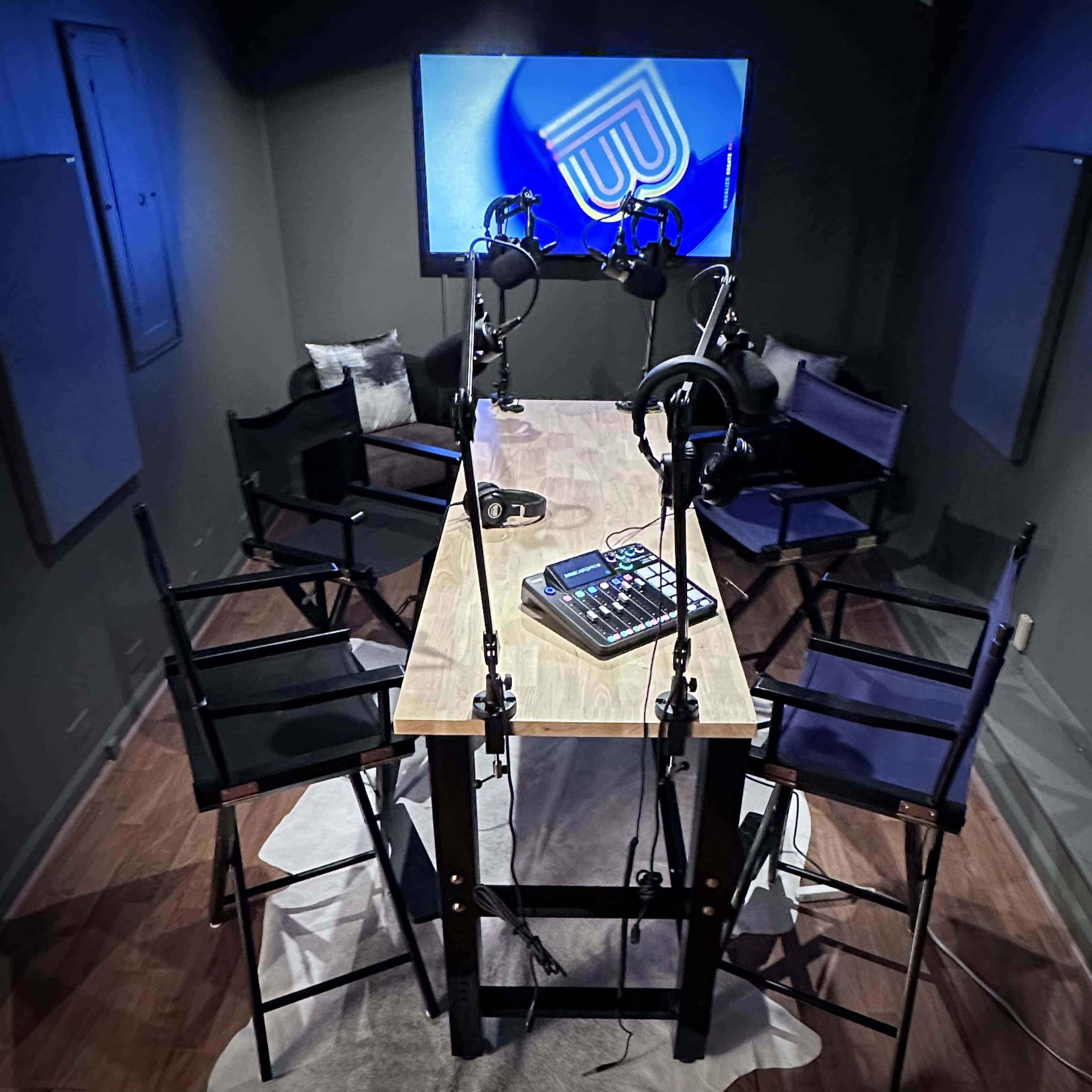The image shows a small recording studio with a large screen, a table in the center, and four microphones positioned for a podcast recording setup.