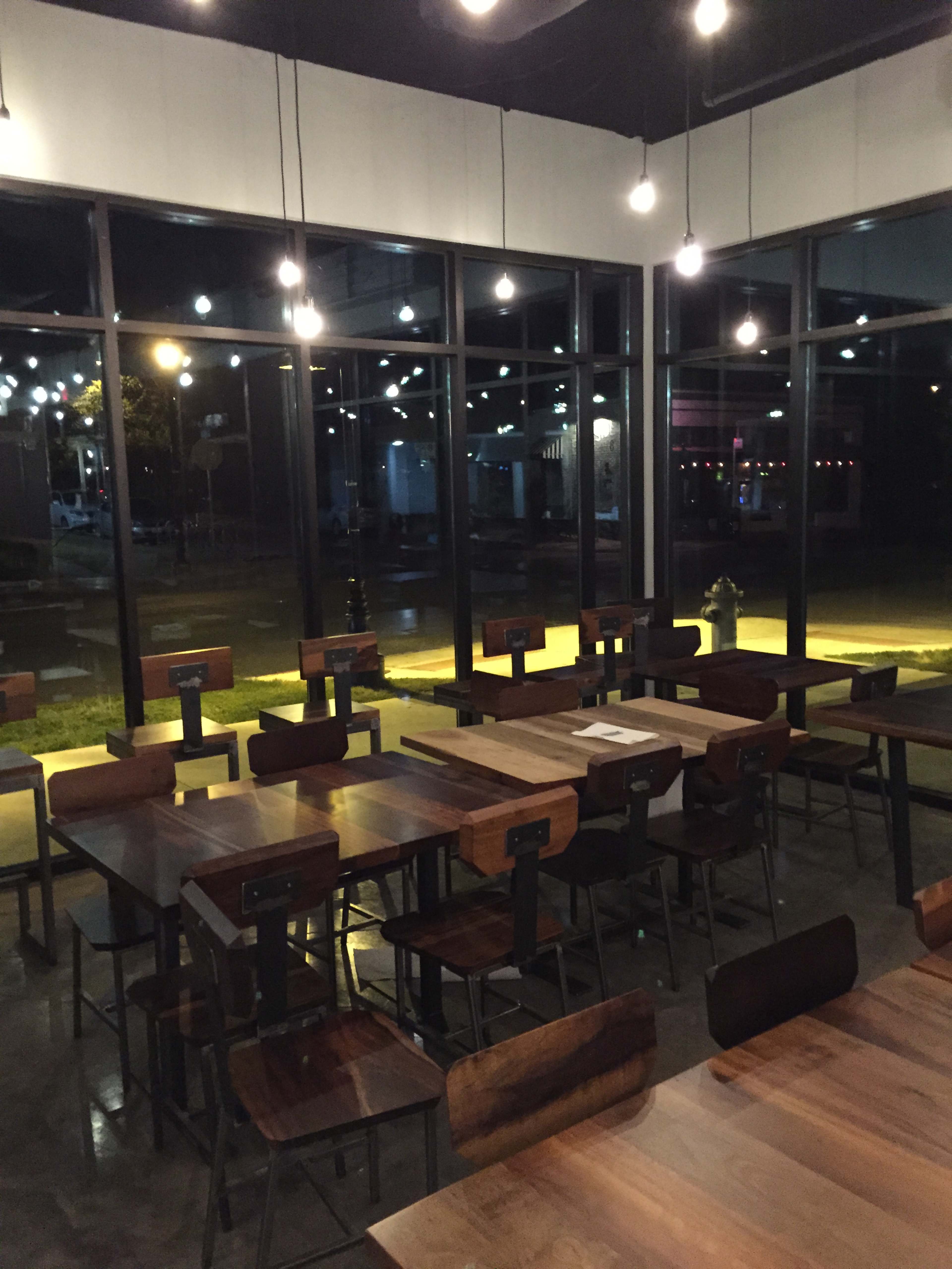 The image depicts a dimly lit restaurant interior with wooden tables and chairs arranged in an open layout, illuminated by hanging light bulbs.