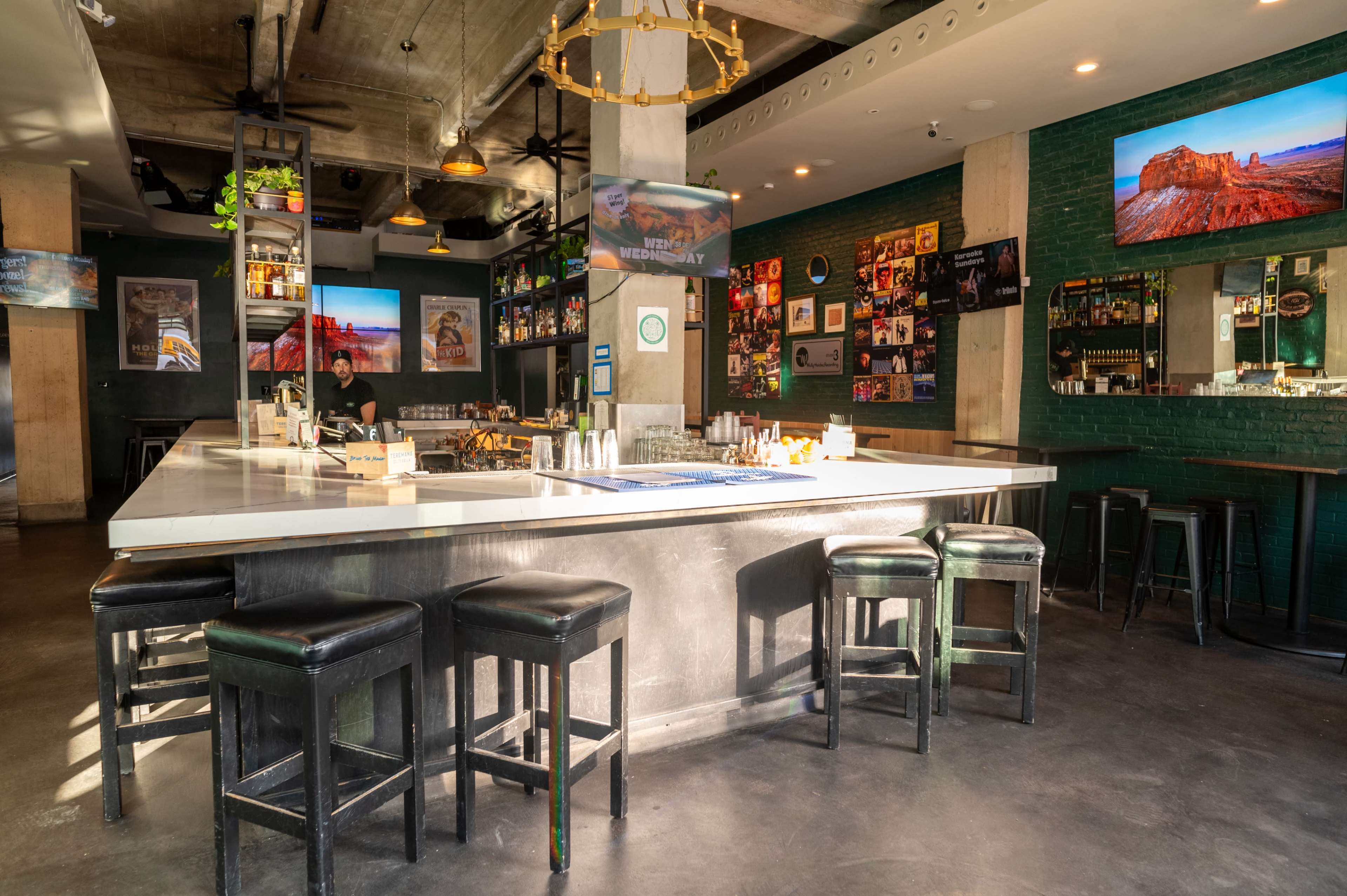 The image shows a modern bar area with a large countertop, black stools, and walls adorned with framed photographs and several screens displaying images.