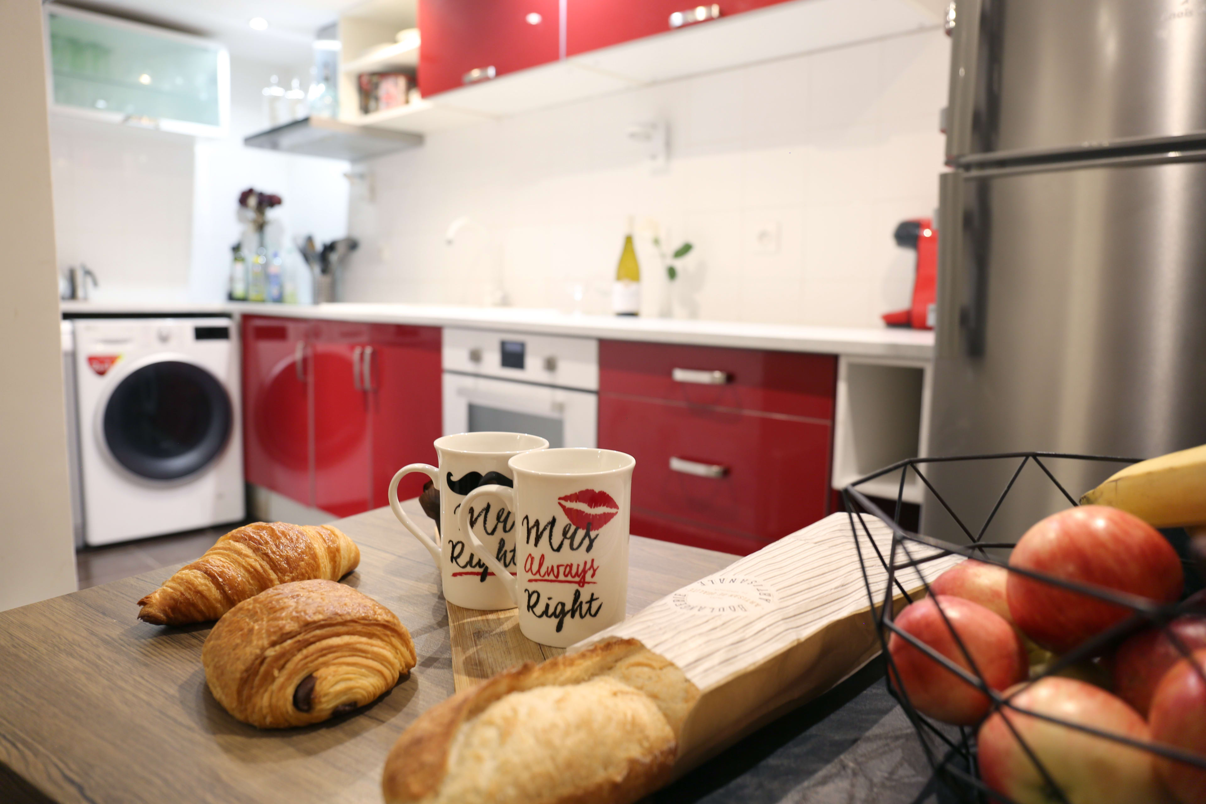 A modern kitchen features a washing machine, red cabinets, two coffee mugs, croissants, and a bowl of apples on a wooden table.