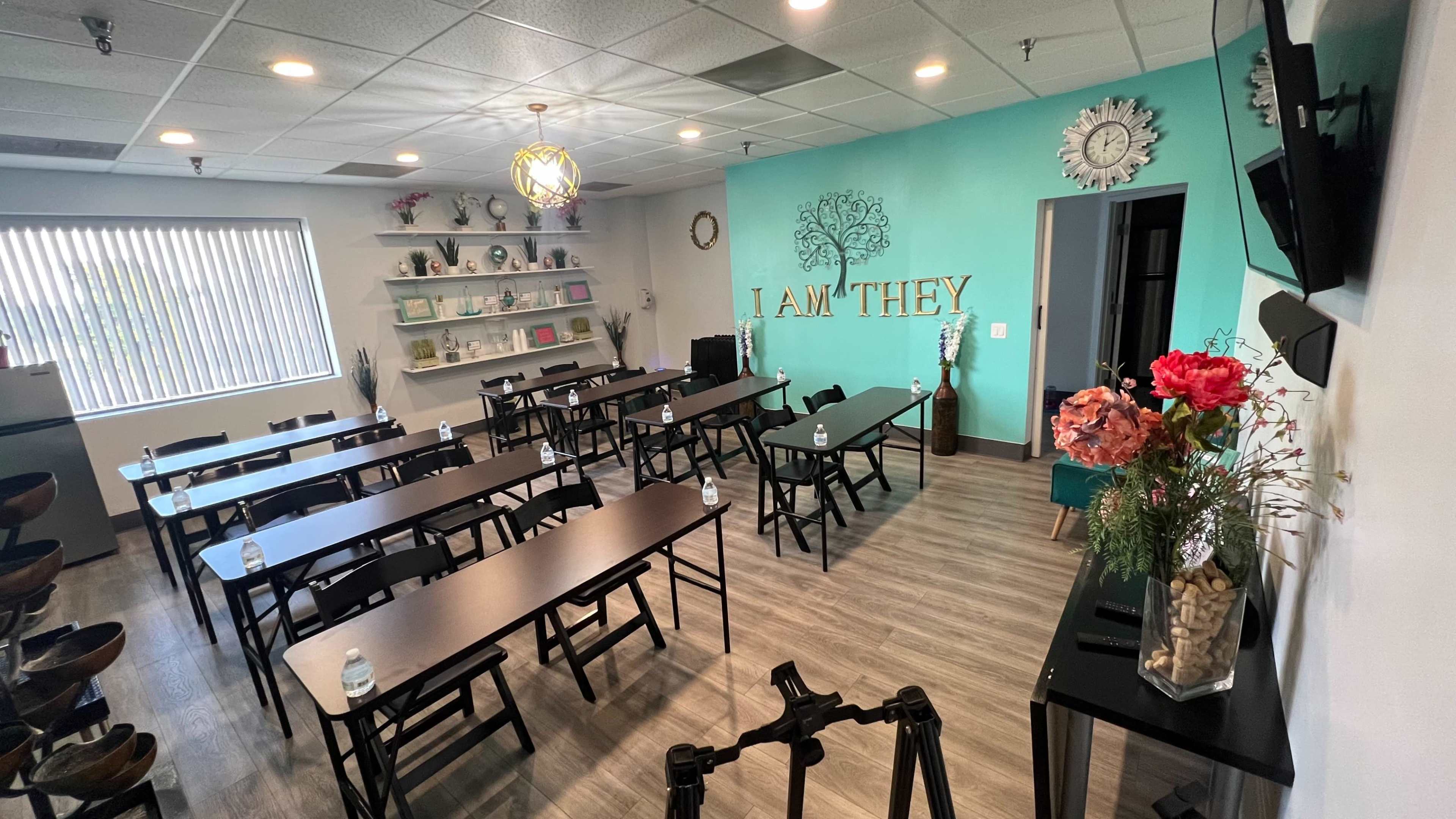 A classroom or meeting space with multiple rows of black tables and chairs, a turquoise wall, and decorative elements including flowers and wall art.