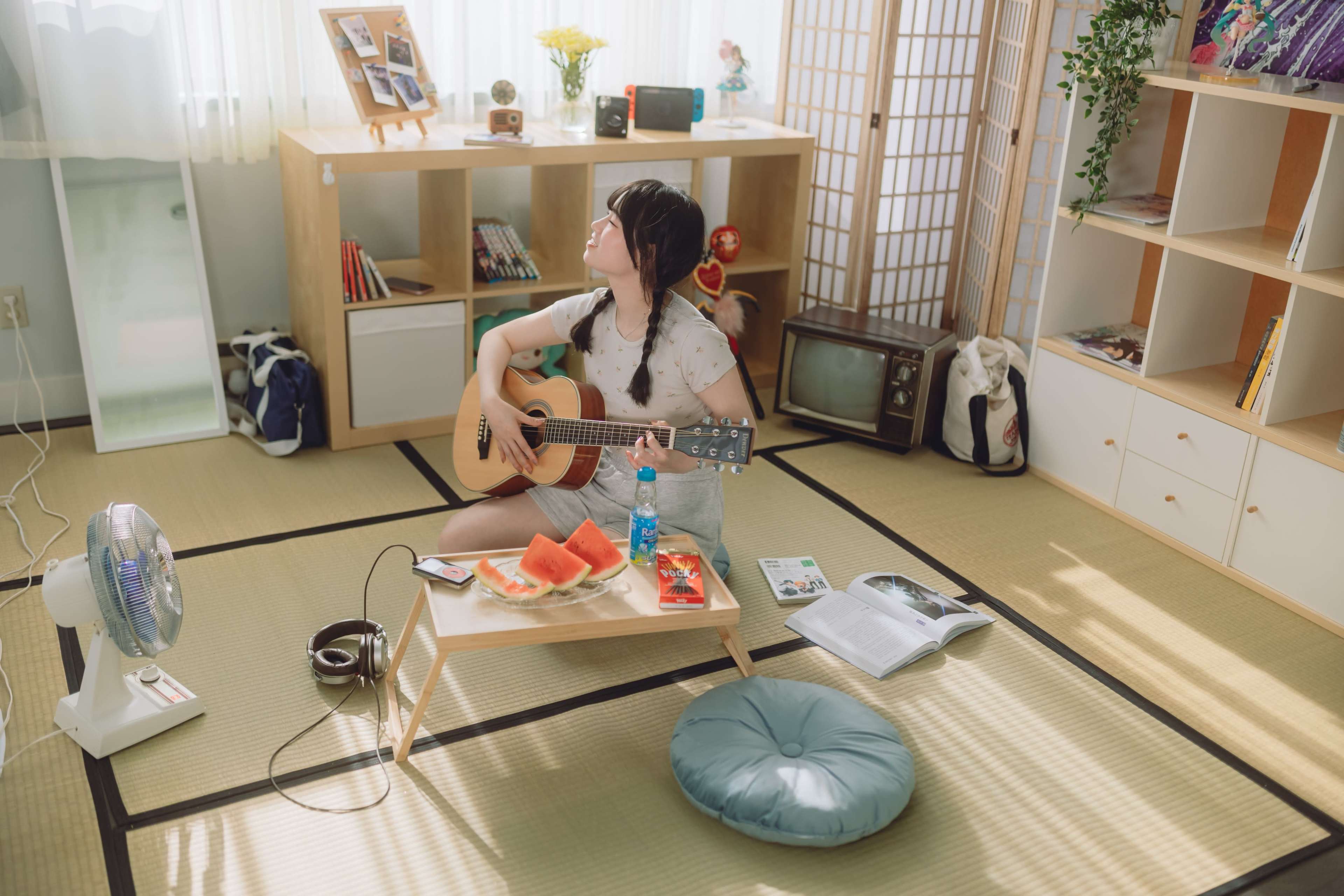 A young woman sits on a floor cushion in a cozy room, playing guitar while surrounded by books, snacks, and a fan.