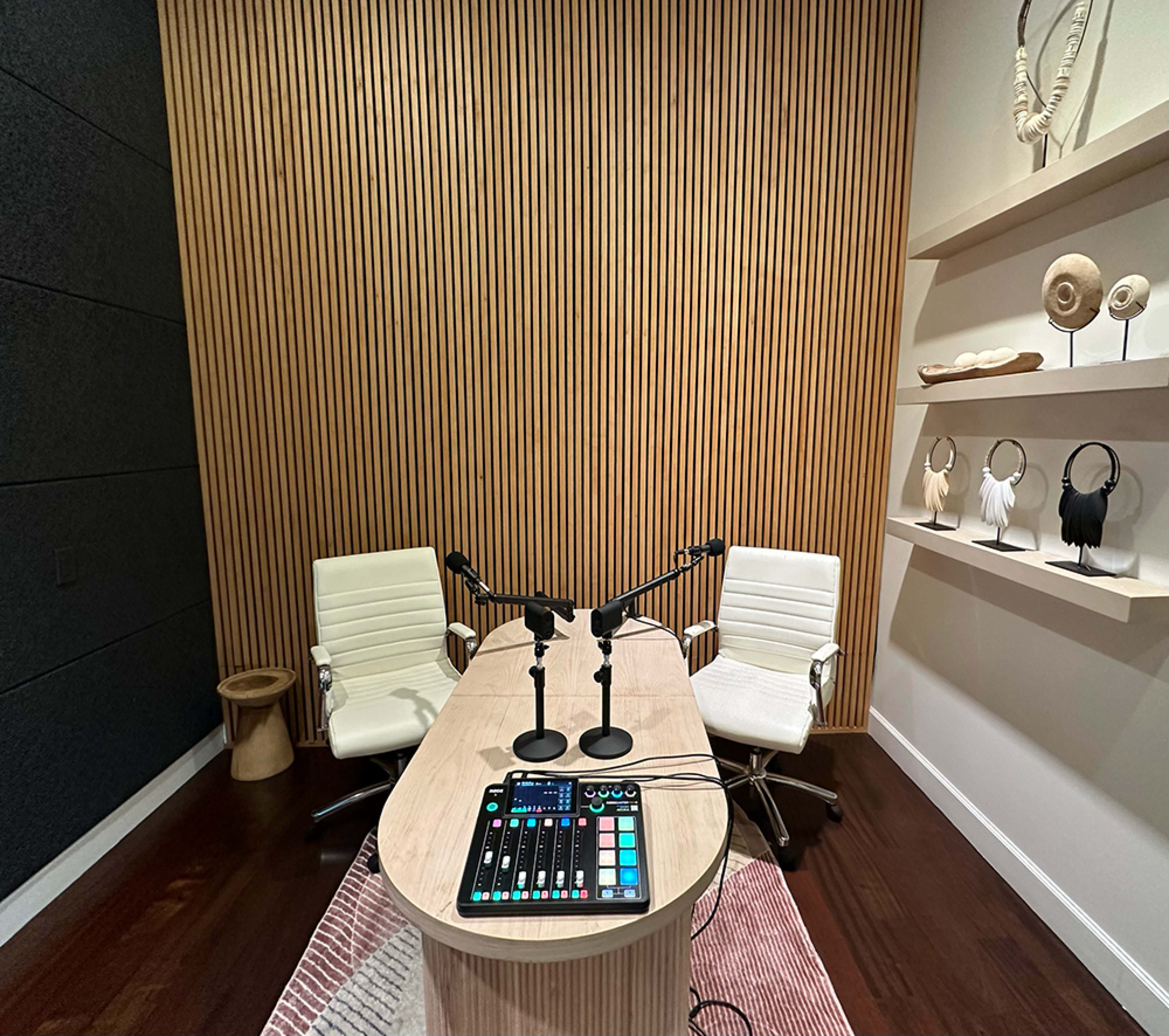 The image shows a small recording studio with two white chairs facing a wooden table equipped with audio mixing equipment, set against a backdrop of slatted wood paneling and decorative shelves.