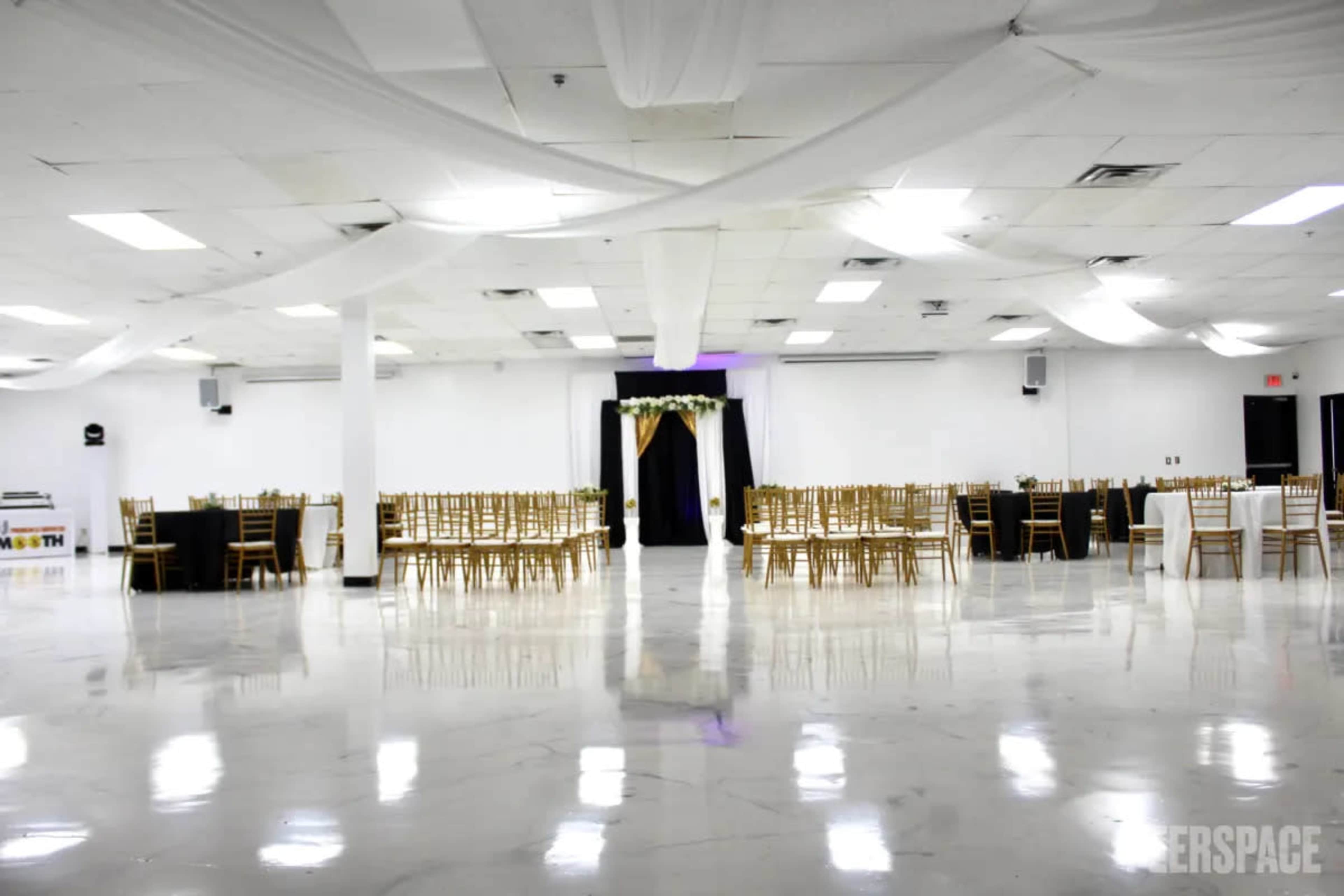 A spacious event hall set up for a gathering, featuring rows of chairs and tables covered in black and gold, with an arch adorned with flowers at one end.