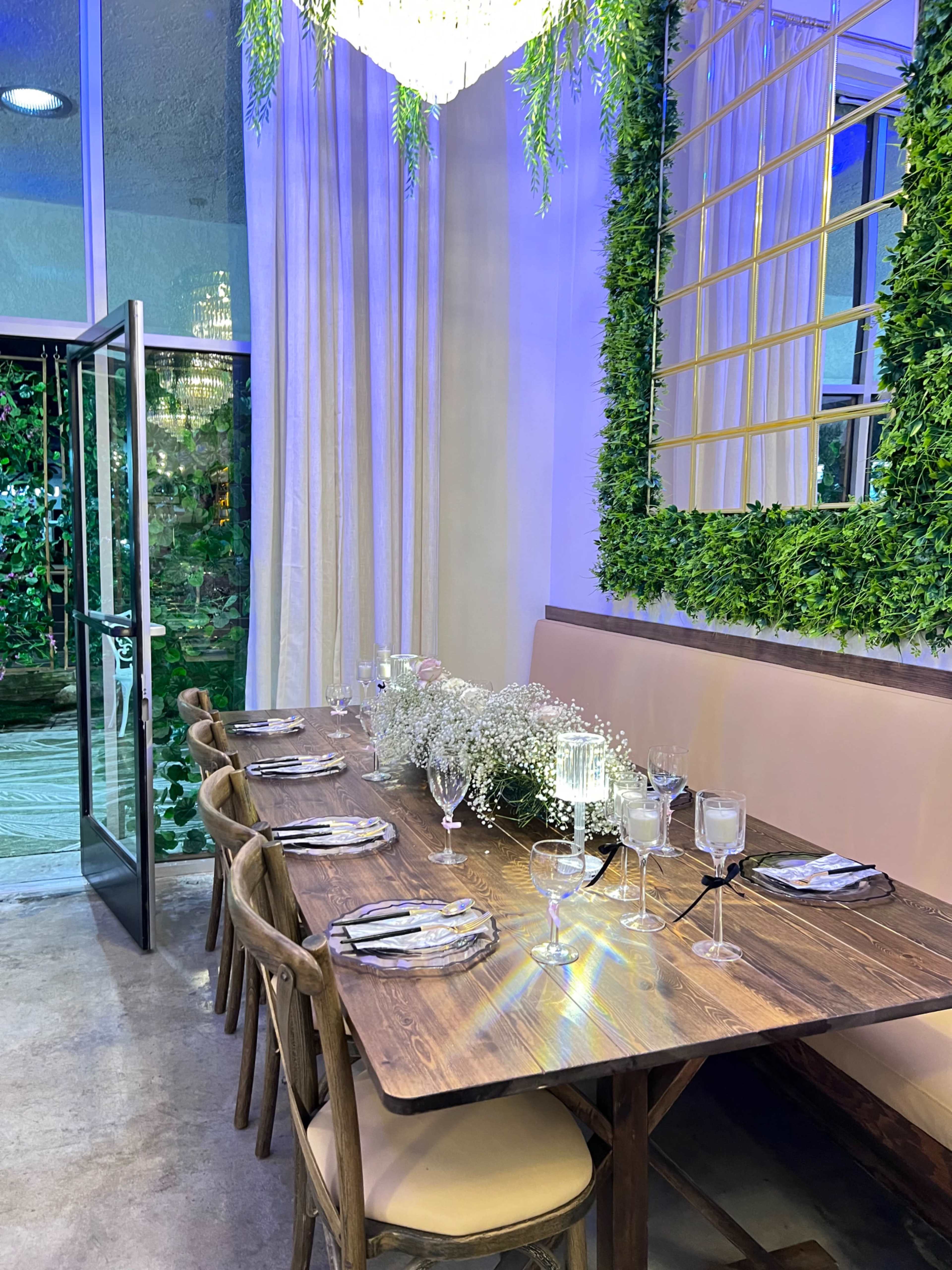 A dining table is set with elegant tableware and a floral centerpiece in a room featuring large windows and greenery accents on the walls.