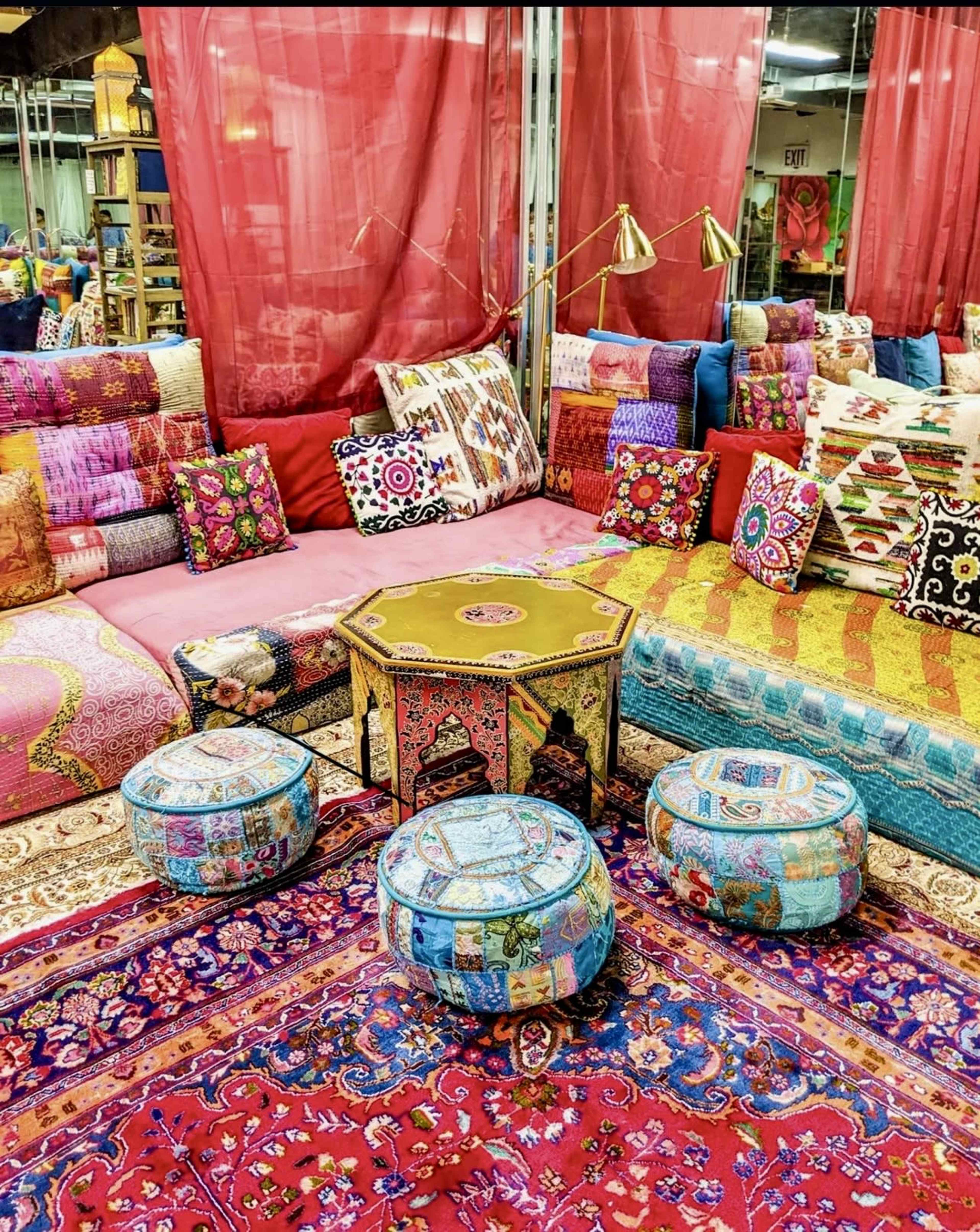 The scene features a colorful arrangement of cushions, pillows, and poufs surrounding a decorative table in an indoor space with vibrant textiles and drapes.
