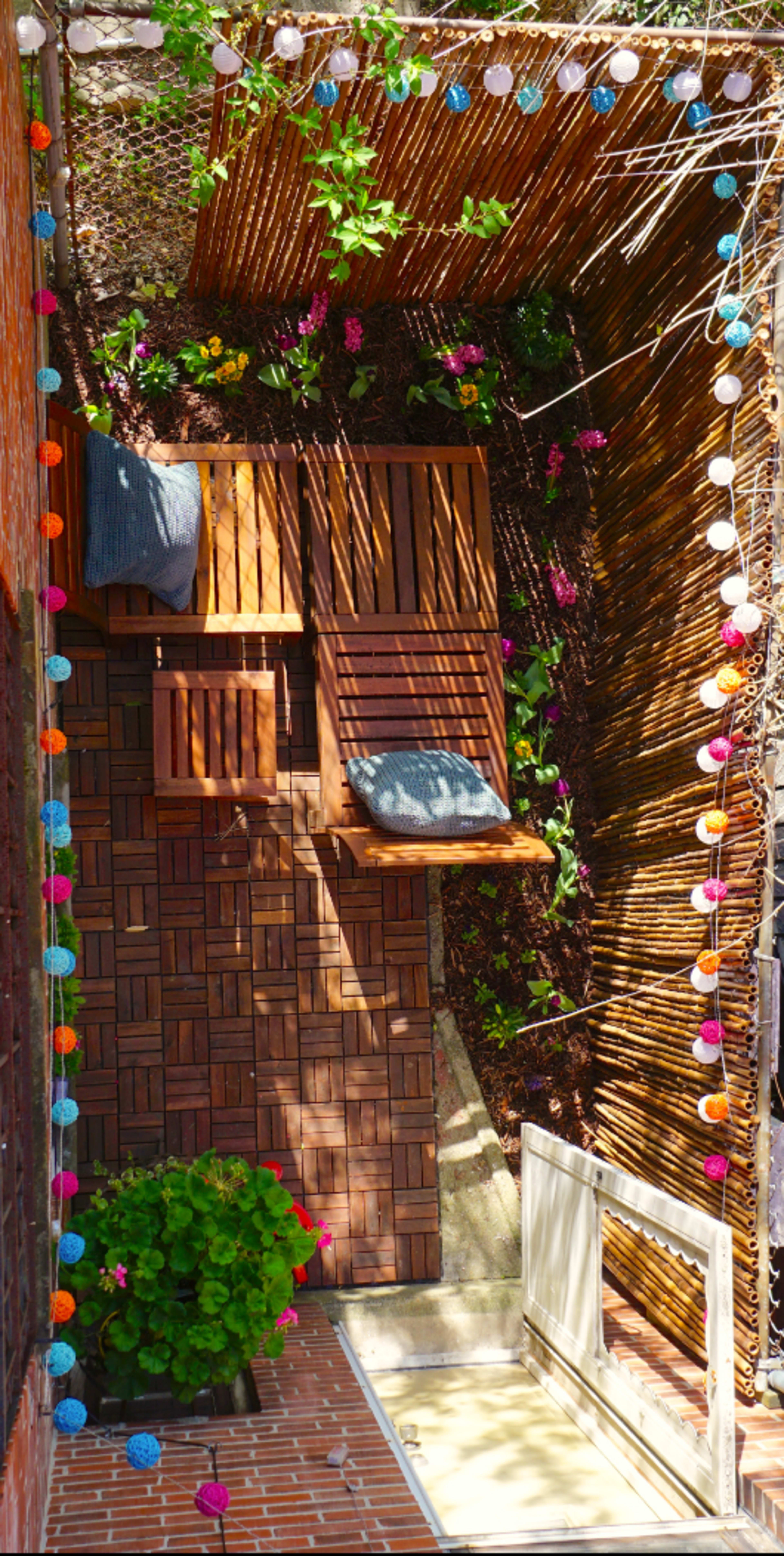 A small outdoor space featuring wooden decking, a cushioned seating area, and colorful string lights along a bamboo fence, surrounded by plants.