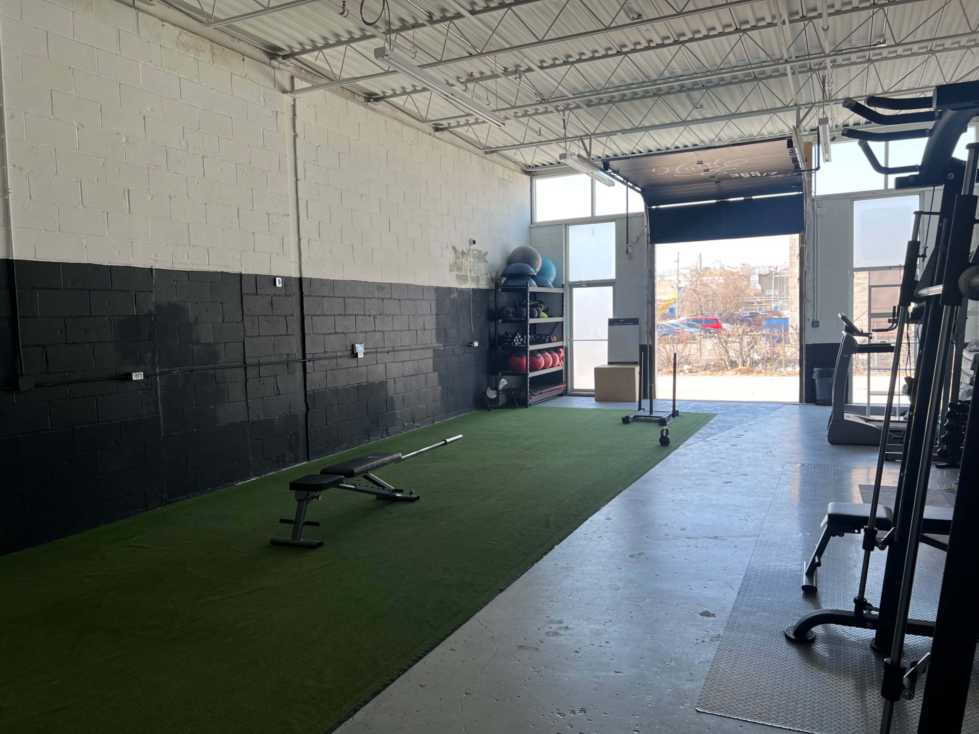 The image shows a gym space featuring a green turf area, weights, and large industrial doors open to the outside.