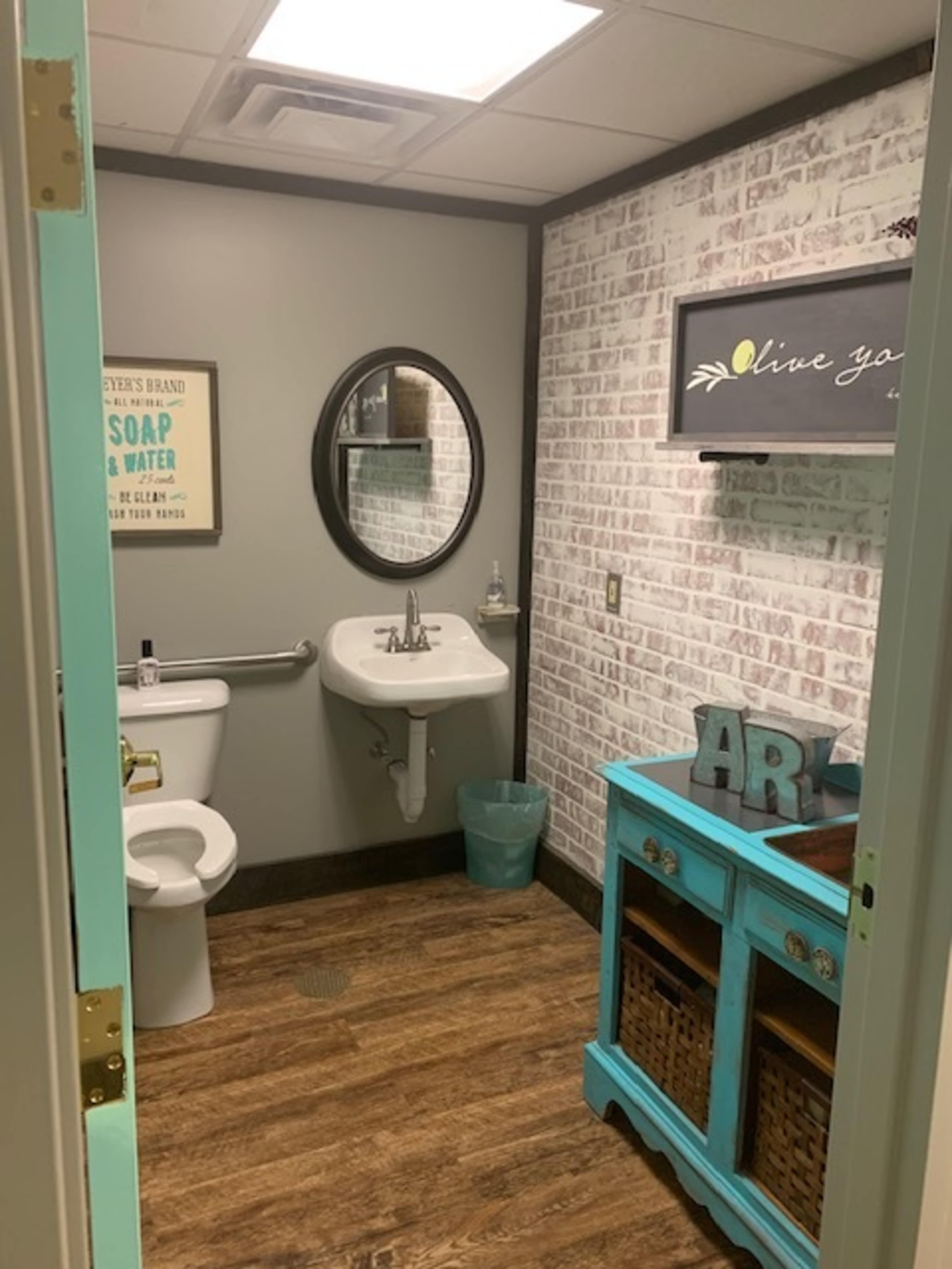 A small bathroom with a toilet, a sink, and a wooden cabinet with baskets, all set against a gray and white brick wall.