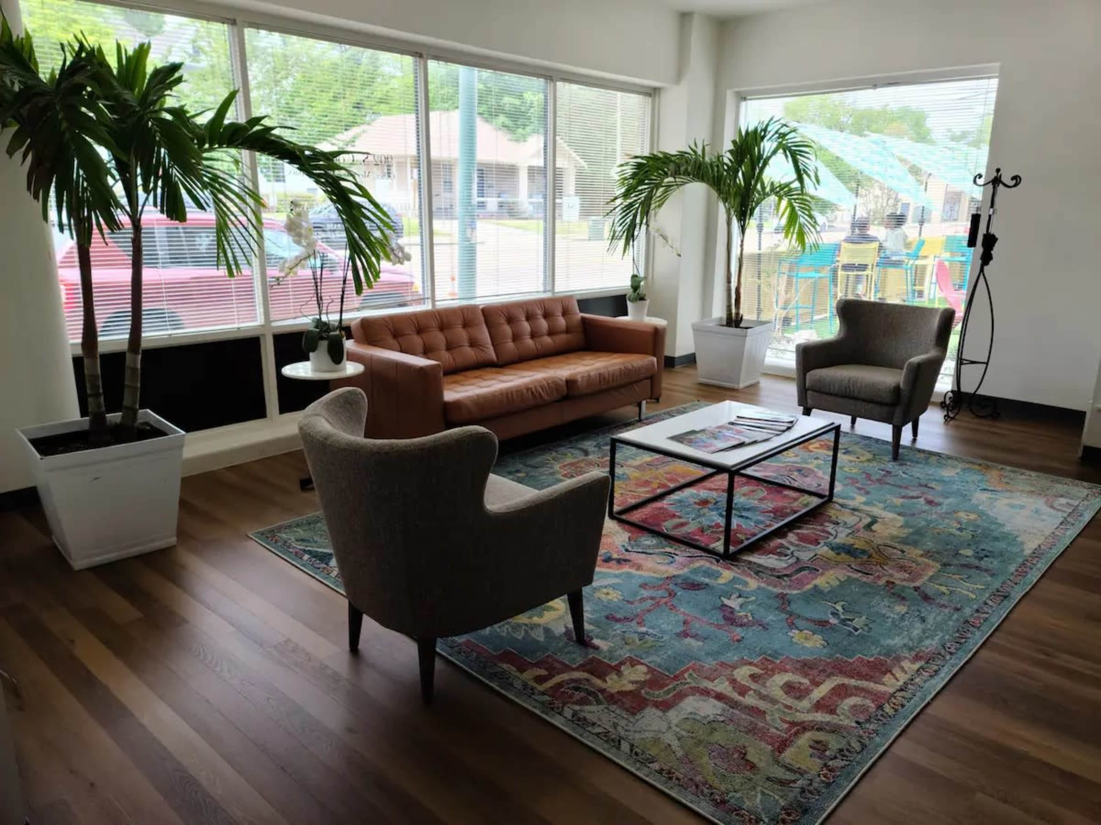 A modern lounge area featuring a brown leather sofa, two upholstered chairs, a coffee table, and tropical plants, with large windows allowing natural light to fill the space.