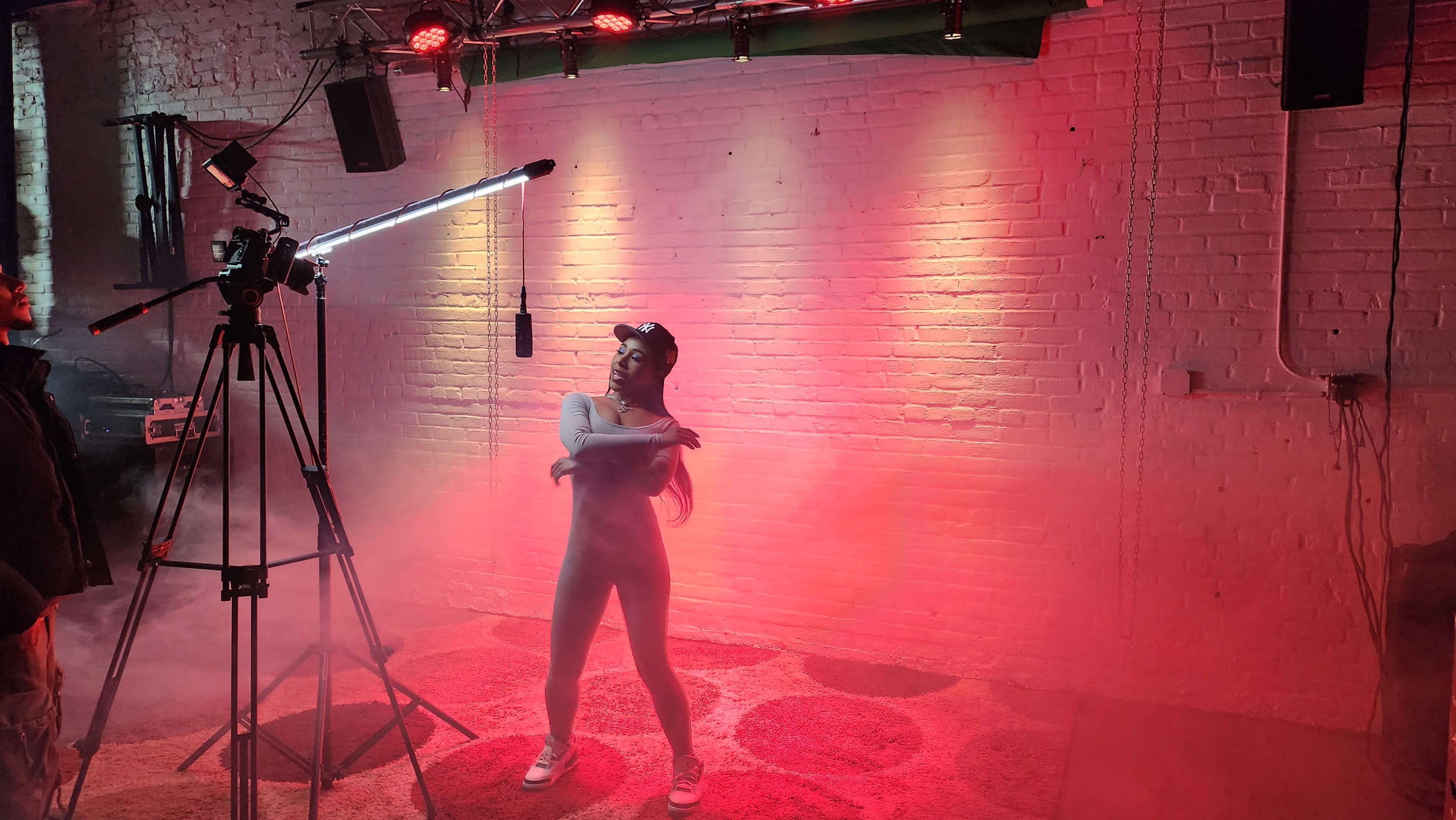 A performer poses in front of a camera setup with colorful lights and fog in a studio setting.