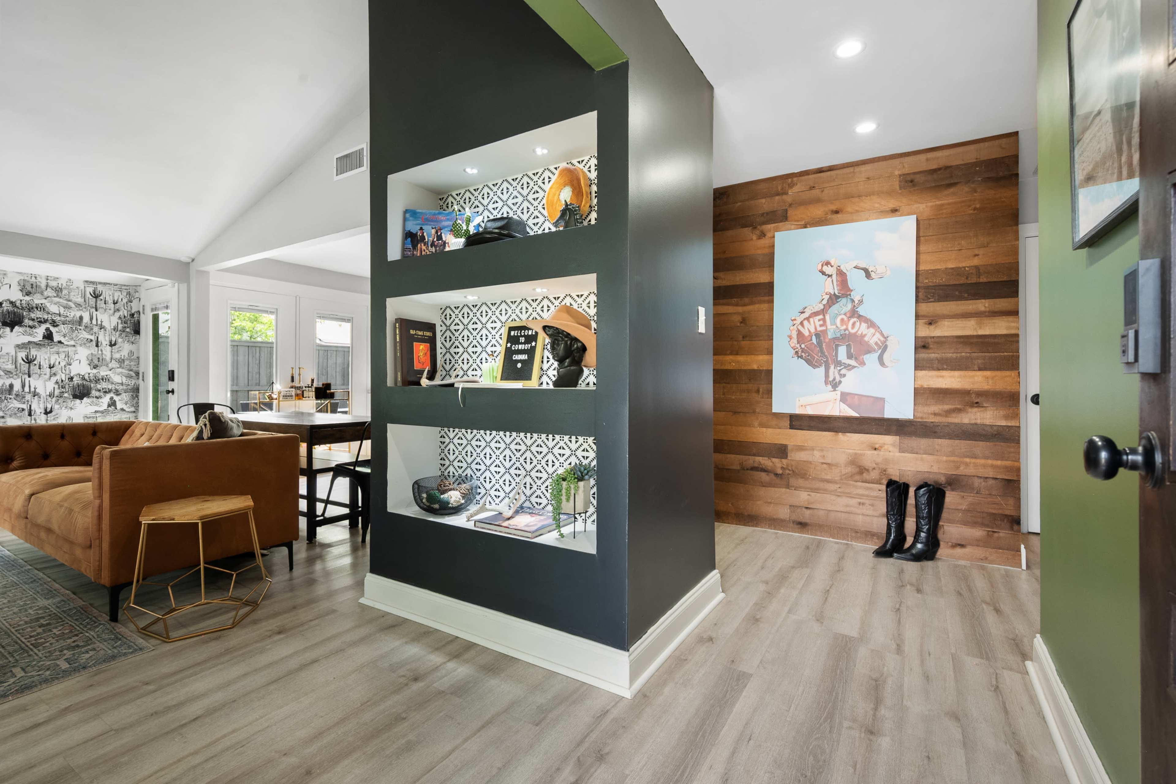 The image shows a modern entryway featuring a dark accent wall with built-in shelves displaying decorative items, a wooden wall with artwork, and a view into a spacious living area with a couch and dining space.