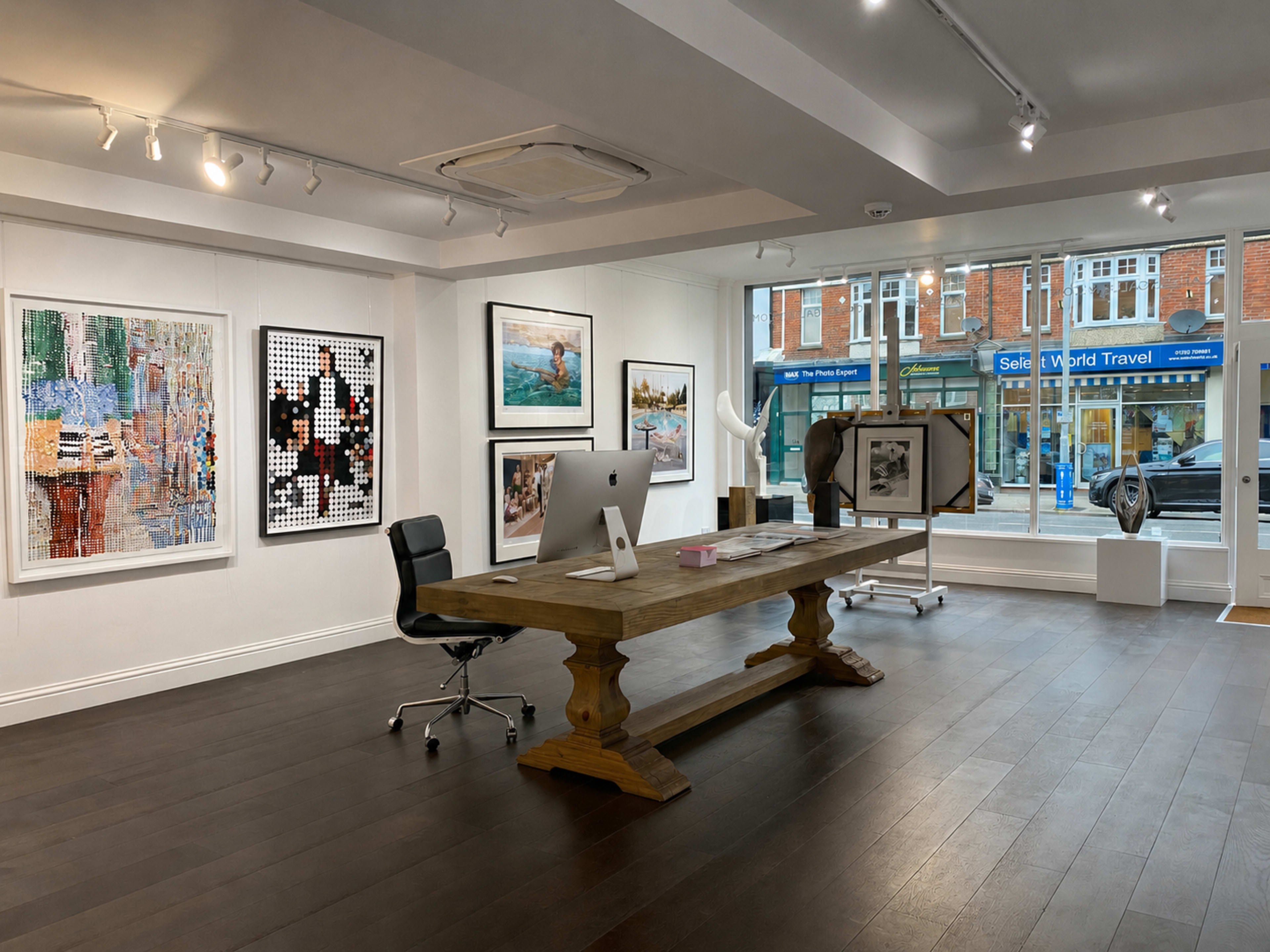 The image shows a modern art gallery featuring a large wooden table, an iMac computer, and various artworks displayed on the walls, with a street view visible through large front windows.