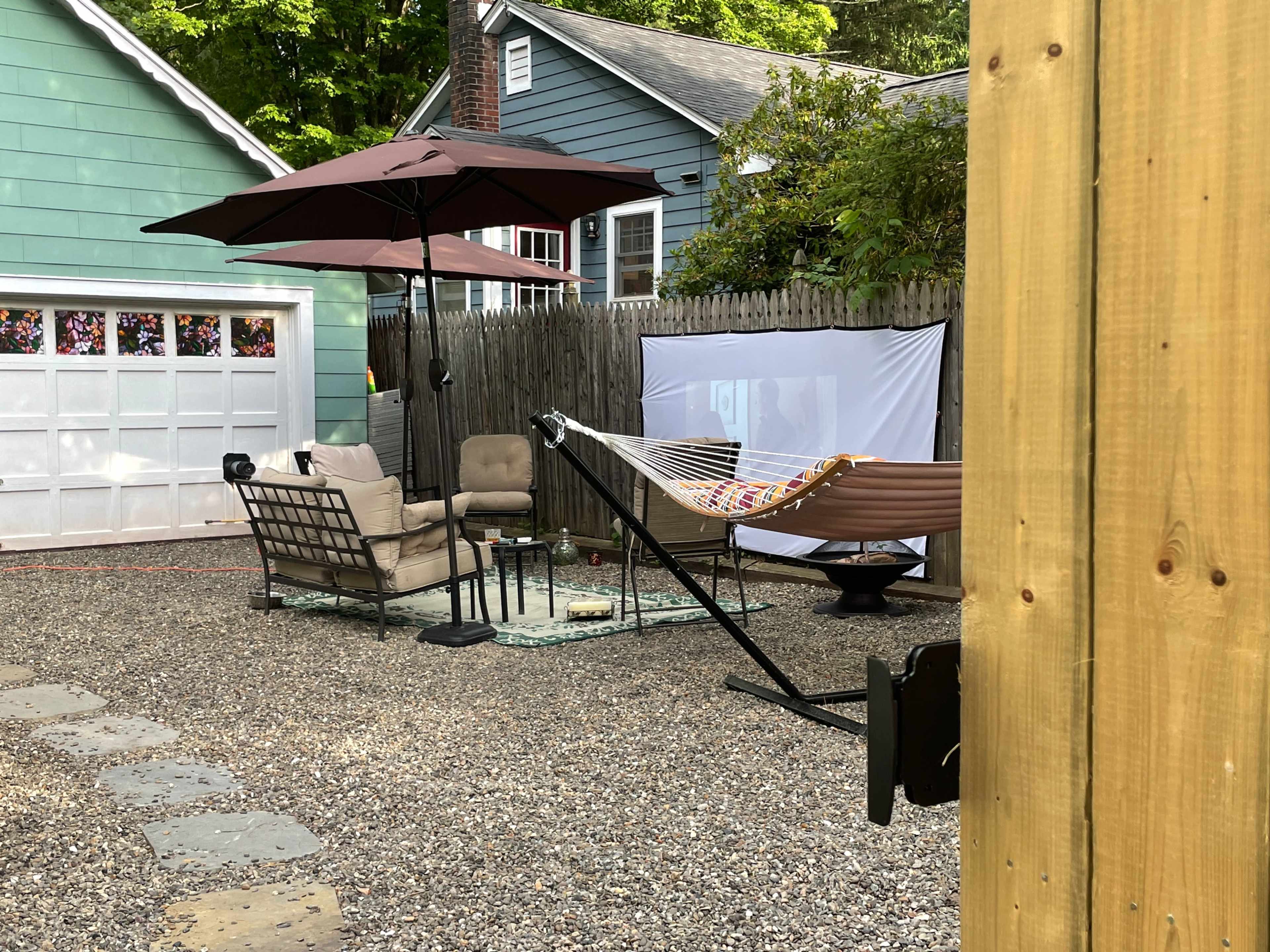 The image shows a backyard space featuring a seating area with a sofa, two umbrellas, a hammock, and a projector screen.