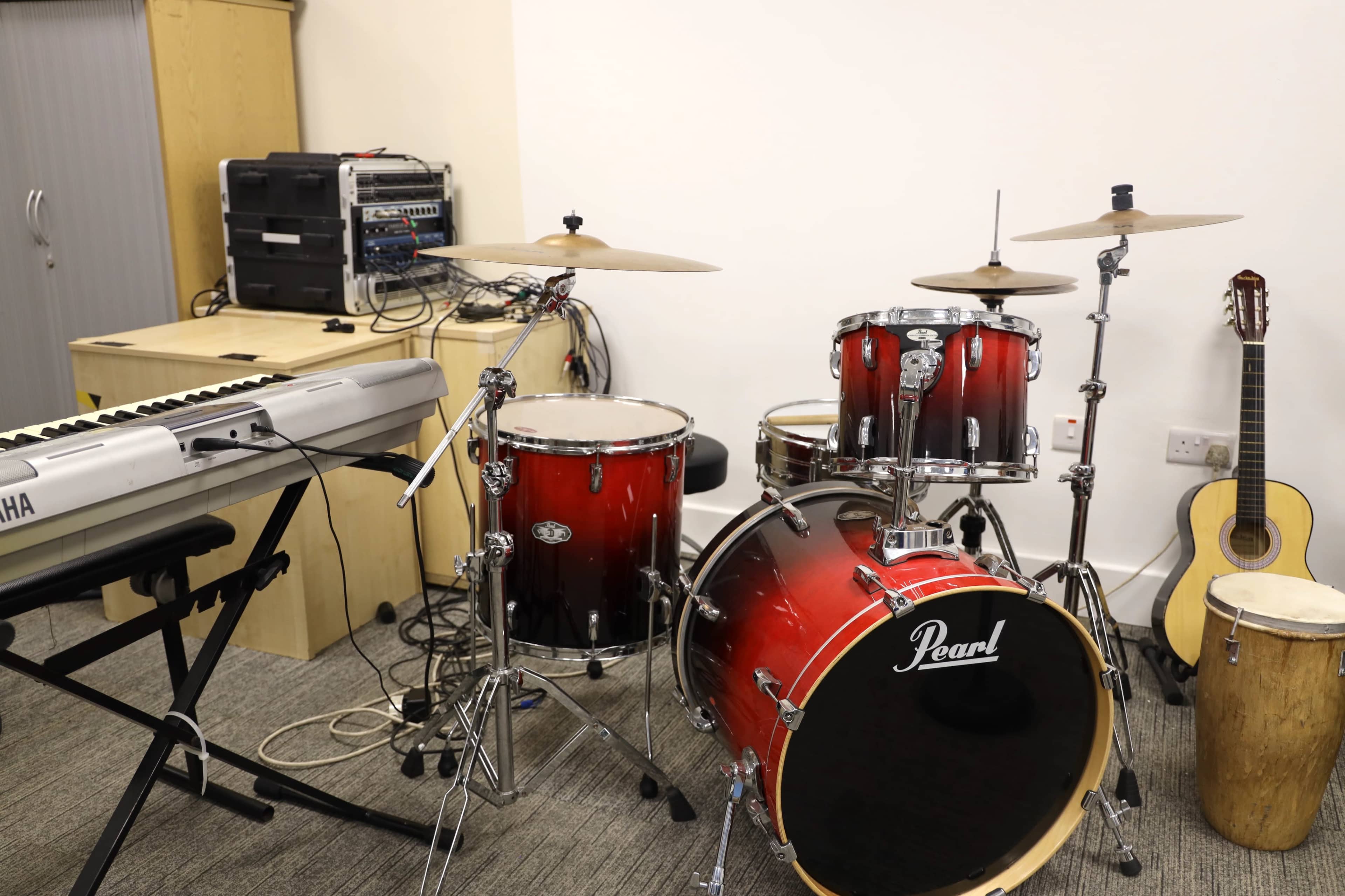The image shows a music practice room containing a red Pearl drum set, a keyboard, a wooden guitar, and various audio equipment on a cabinet.