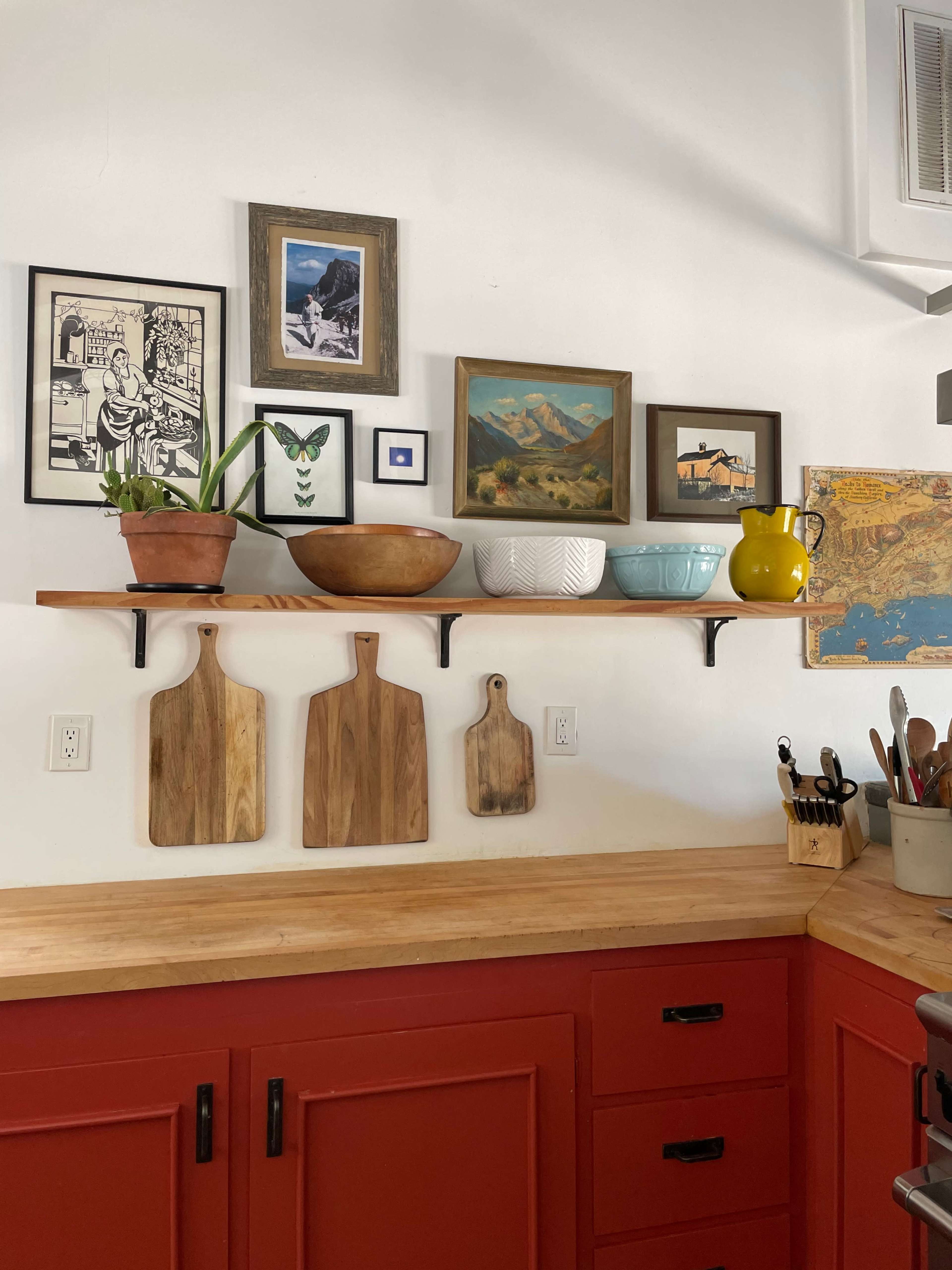 The image shows a kitchen wall with a wooden shelf holding various decorative items, including framed artwork, a potted plant, ceramic bowls, and wooden cutting boards.