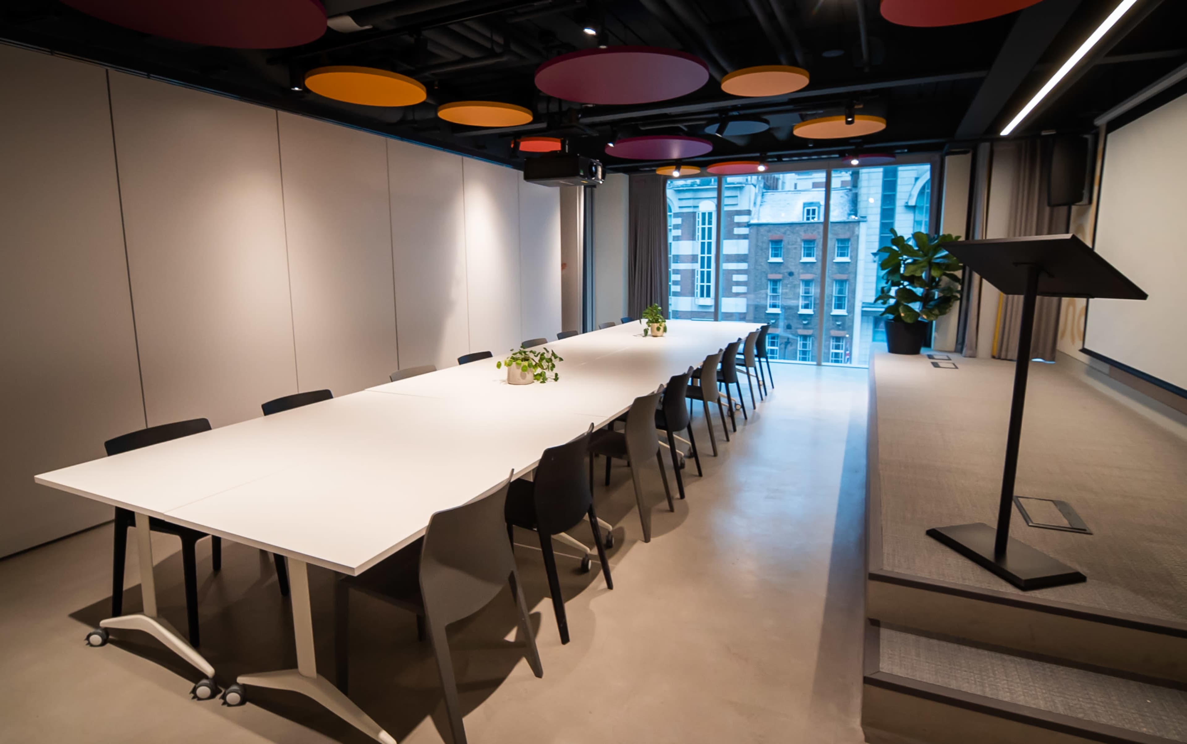 A conference room features a long white table surrounded by black chairs, with colorful circular ceiling panels and a large window overlooking a cityscape.