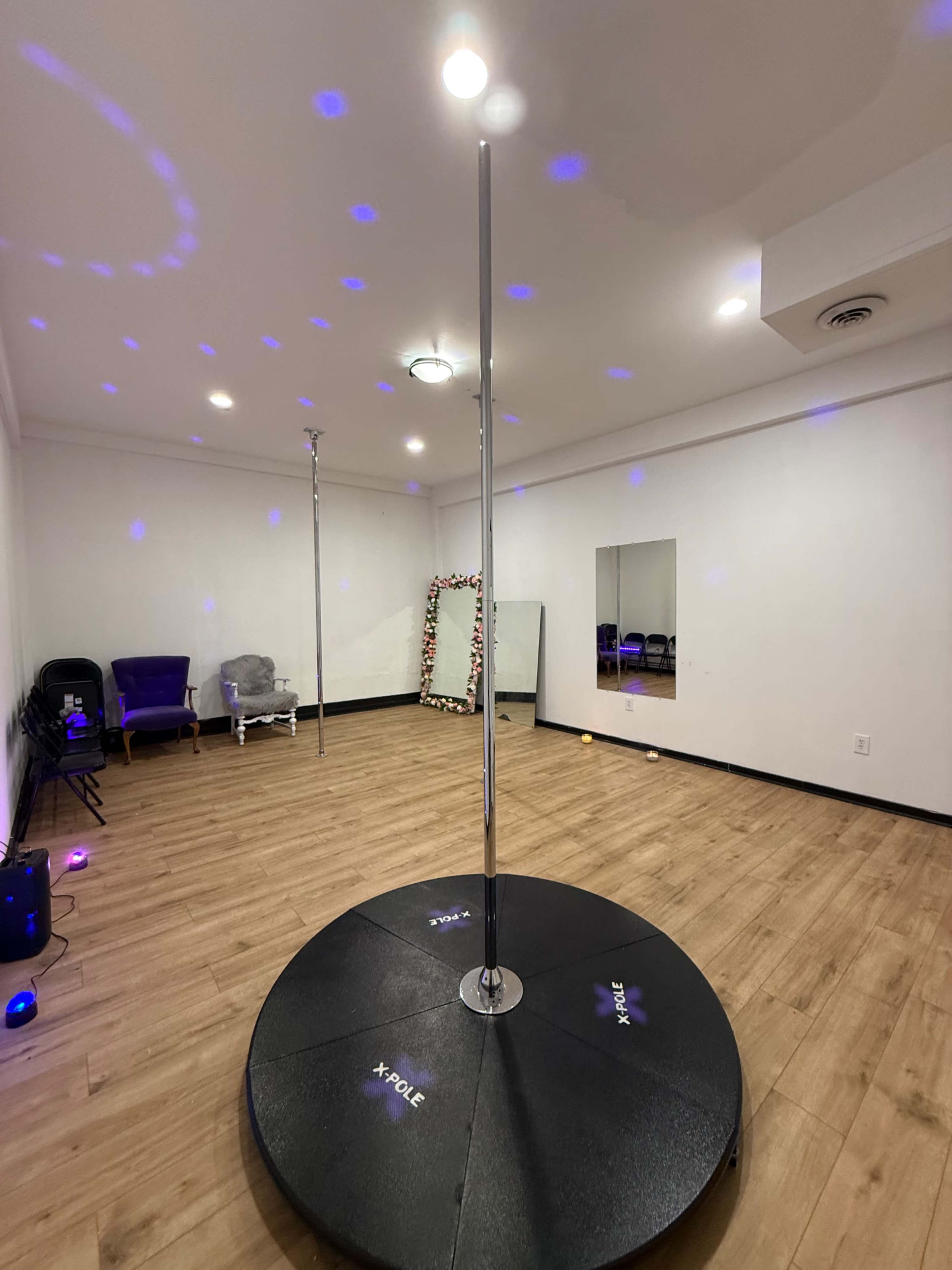 The image shows an empty pole dance studio with a single chrome pole on a circular black platform, surrounded by mirrors and dim lighting.