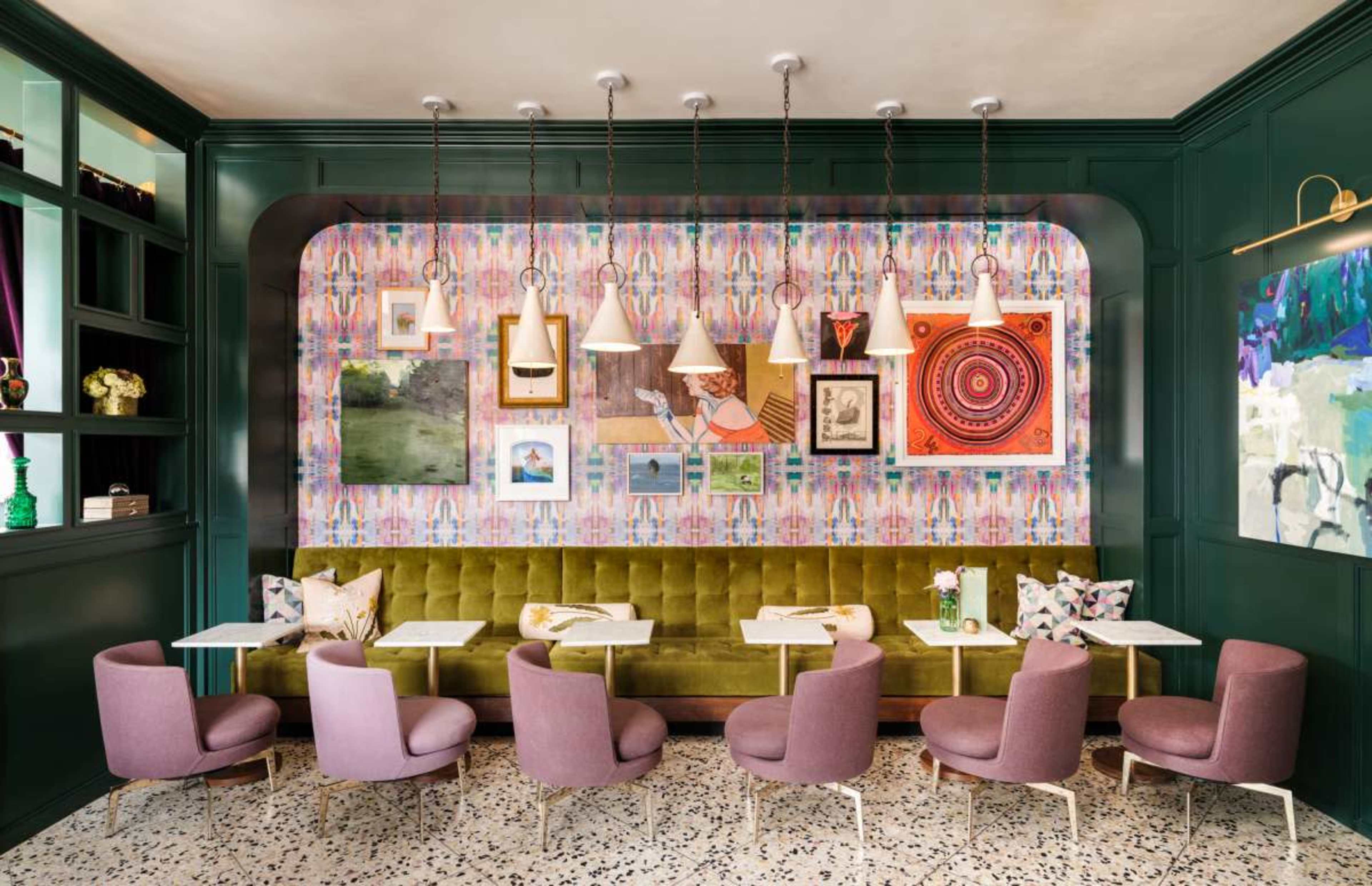 The image shows a modern café interior with a green wall featuring a colorful gallery of framed art, a long green banquette, and purple upholstered chairs arranged around small white tables.