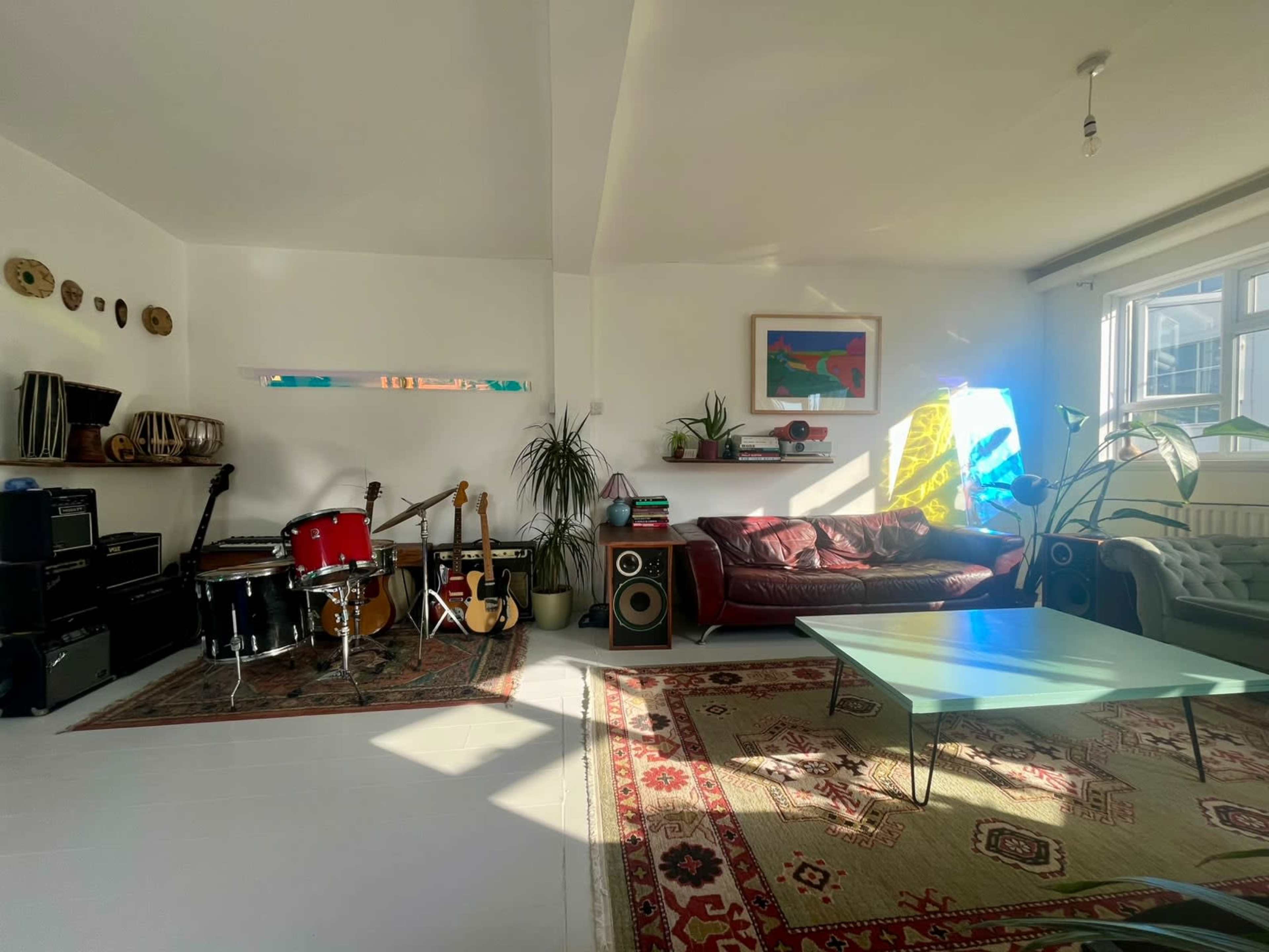 The image shows a bright living room featuring musical instruments, a red drum set, various guitars, a large sofa, a glass coffee table, and colorful artwork on the walls.