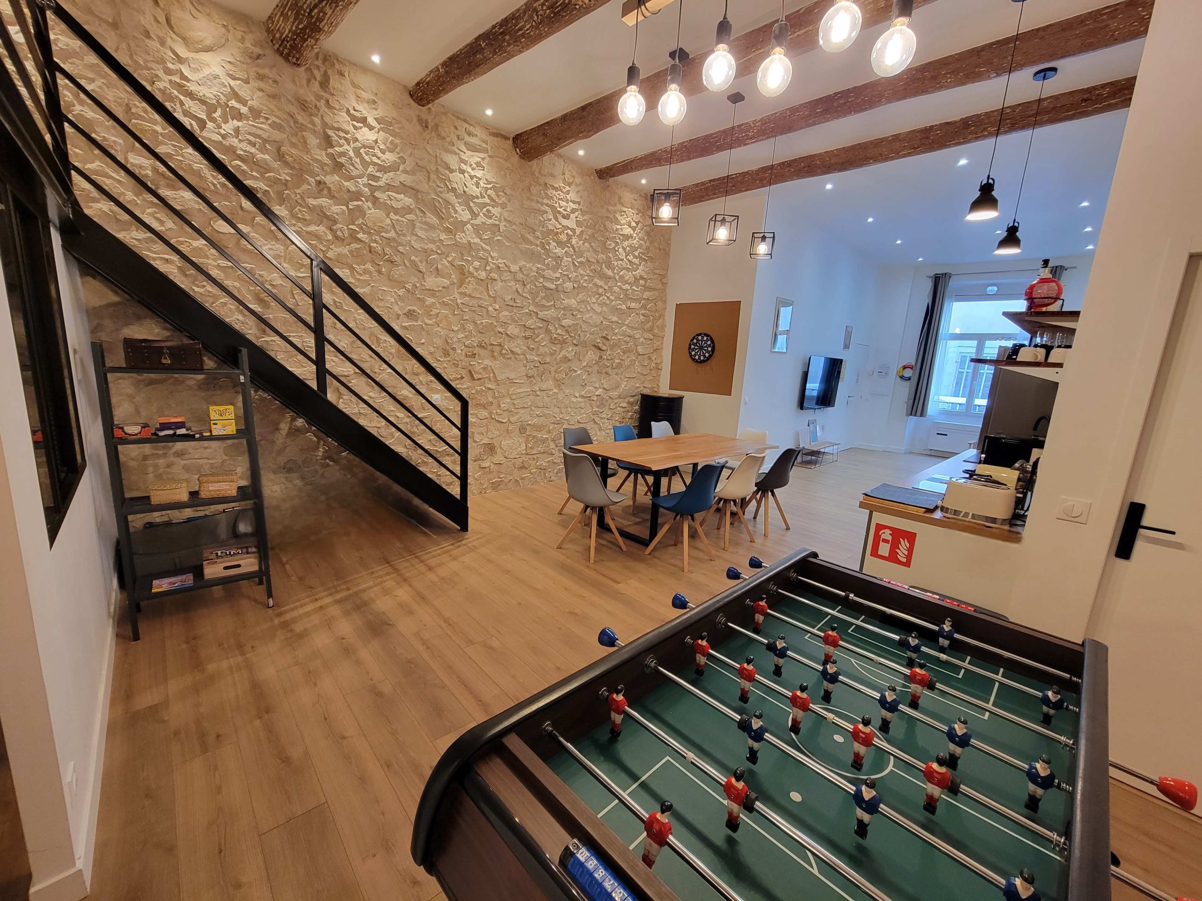 The image shows a modern living space featuring a foosball table in the foreground, a dining area with a round table, and a staircase leading to a loft.