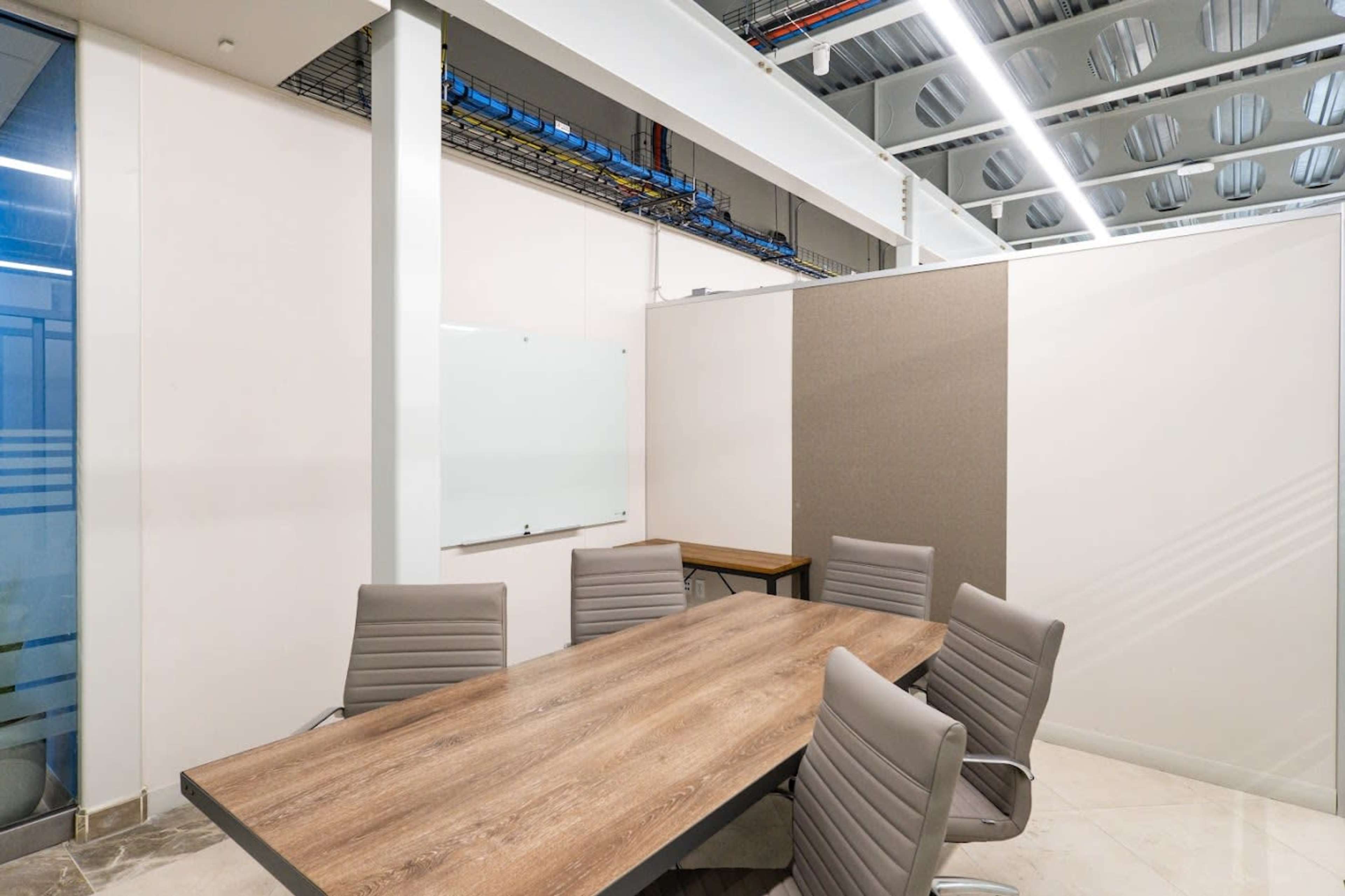 The image shows a modern meeting room featuring a wooden conference table surrounded by six gray chairs, with a whiteboard and a partially partitioned wall in the background.