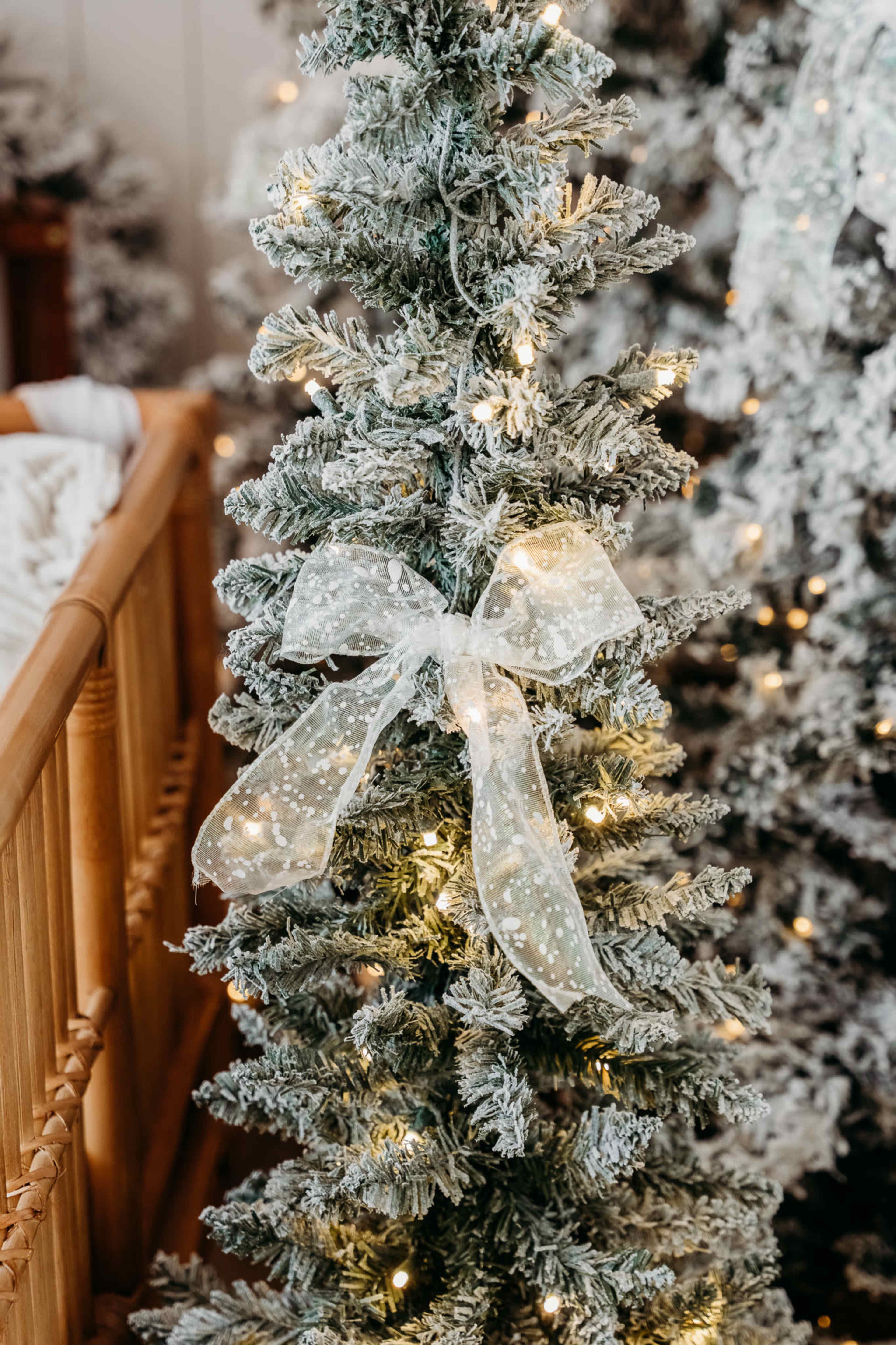 A frosted Christmas tree decorated with white lights and a sheer ribbon bow stands next to a wooden crib in a cozy room.