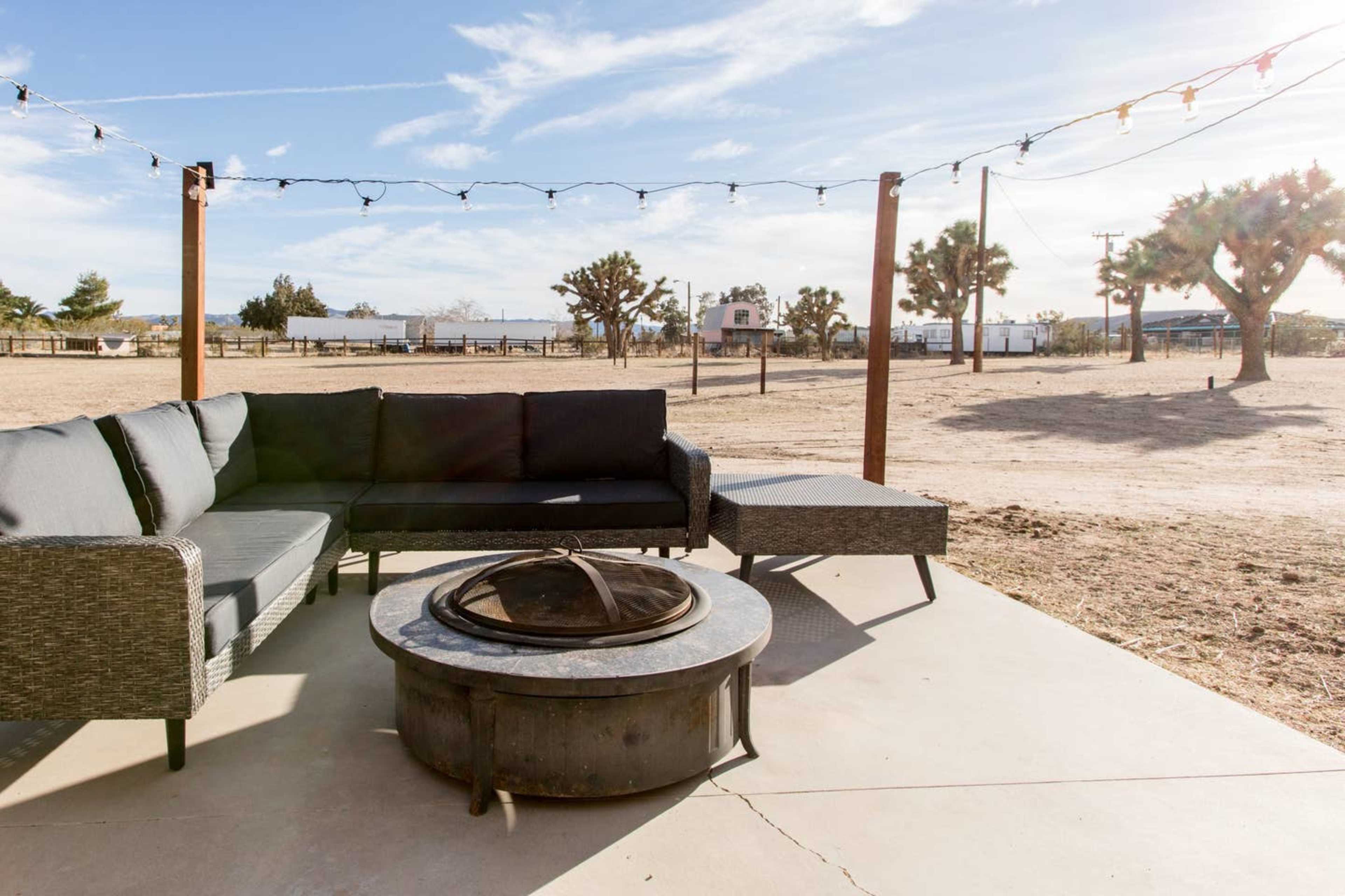 A patio area features a sectional sofa and circular fire pit, surrounded by a desert landscape with sparse vegetation and distant structures.