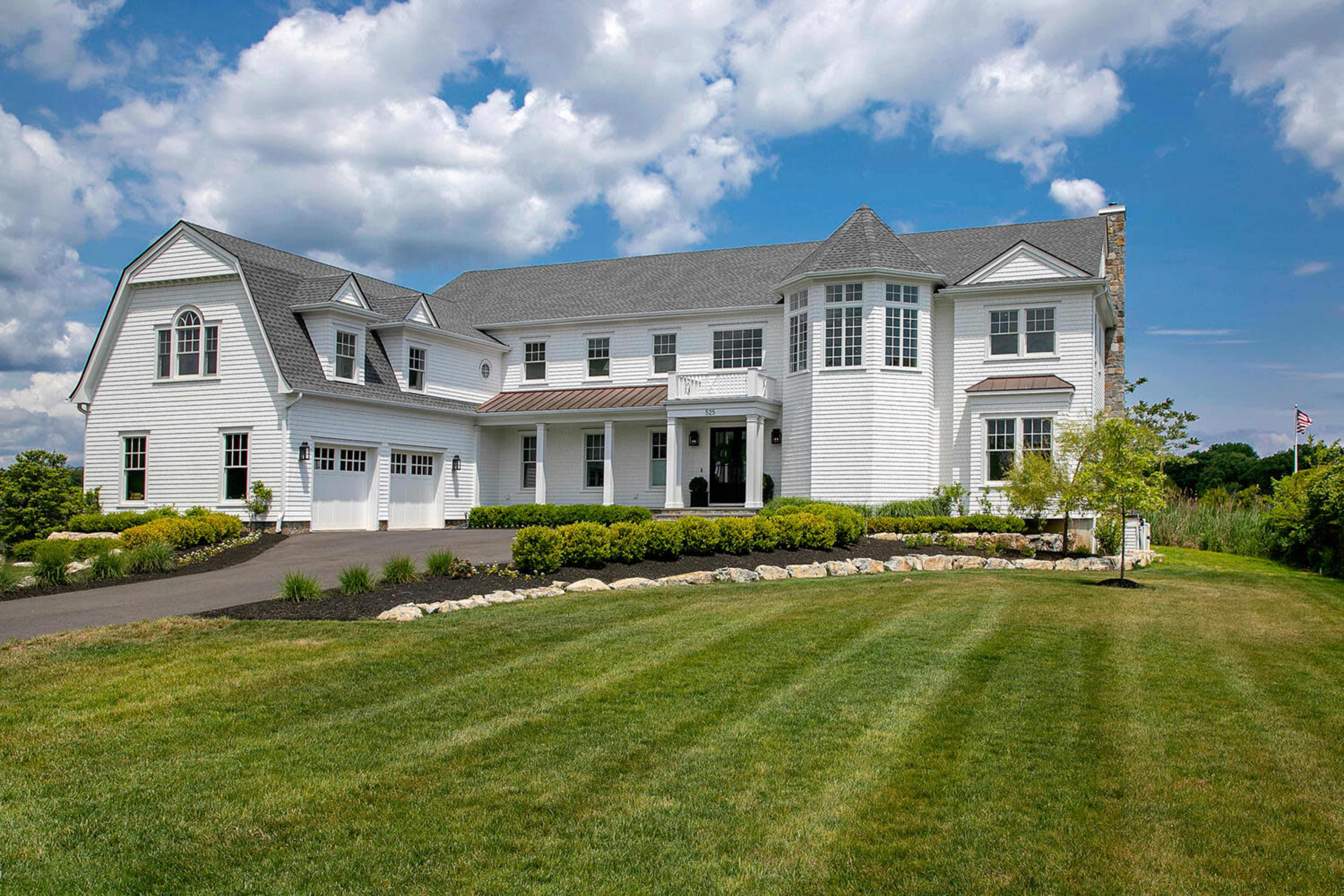 A large white house with a gray roof, featuring a front porch, a landscaped yard, and a paved driveway.
