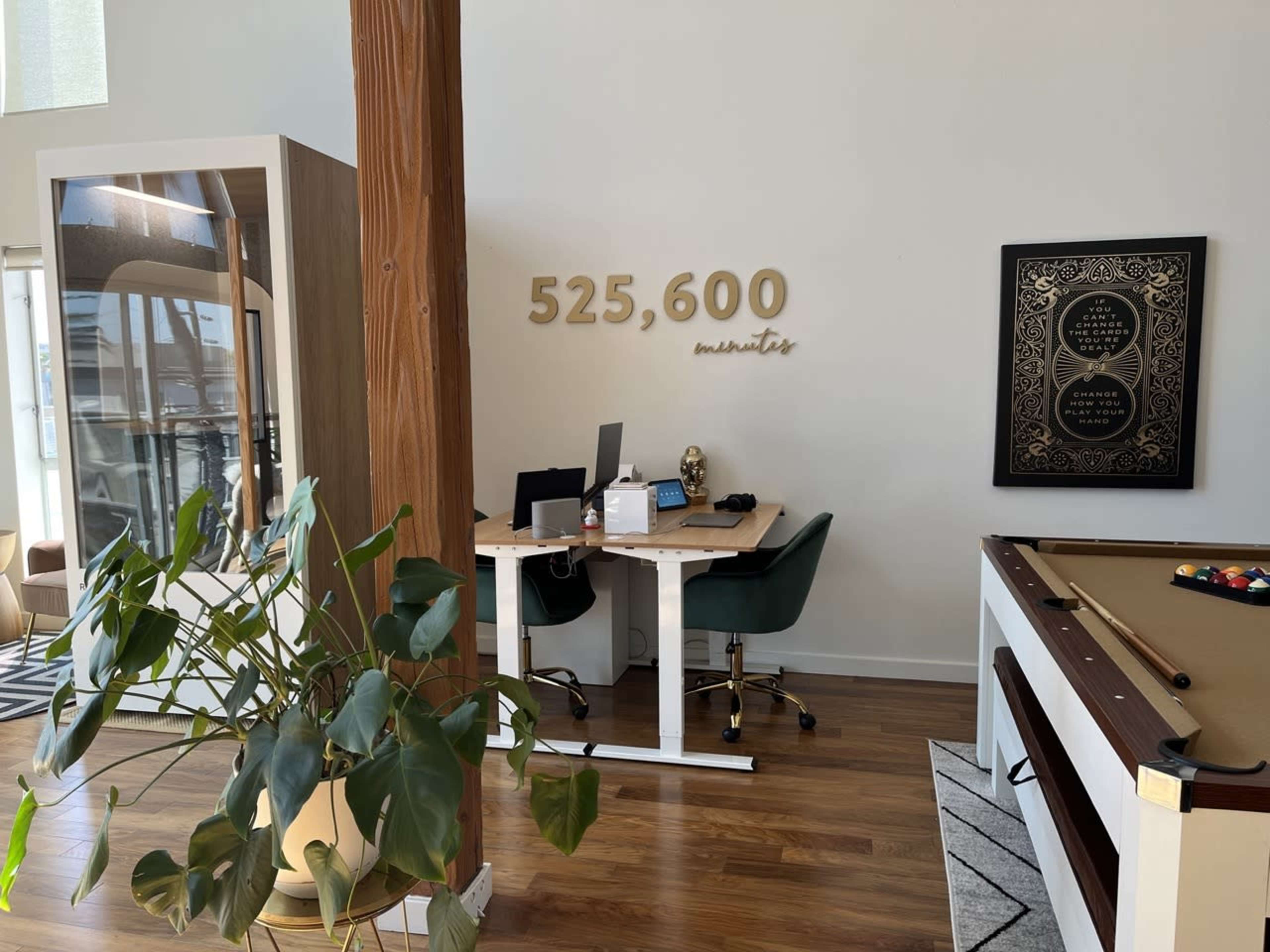 A modern office space featuring a desk with a computer, a green chair, a decorative wall with "525,600 minutes," and a pool table nearby.
