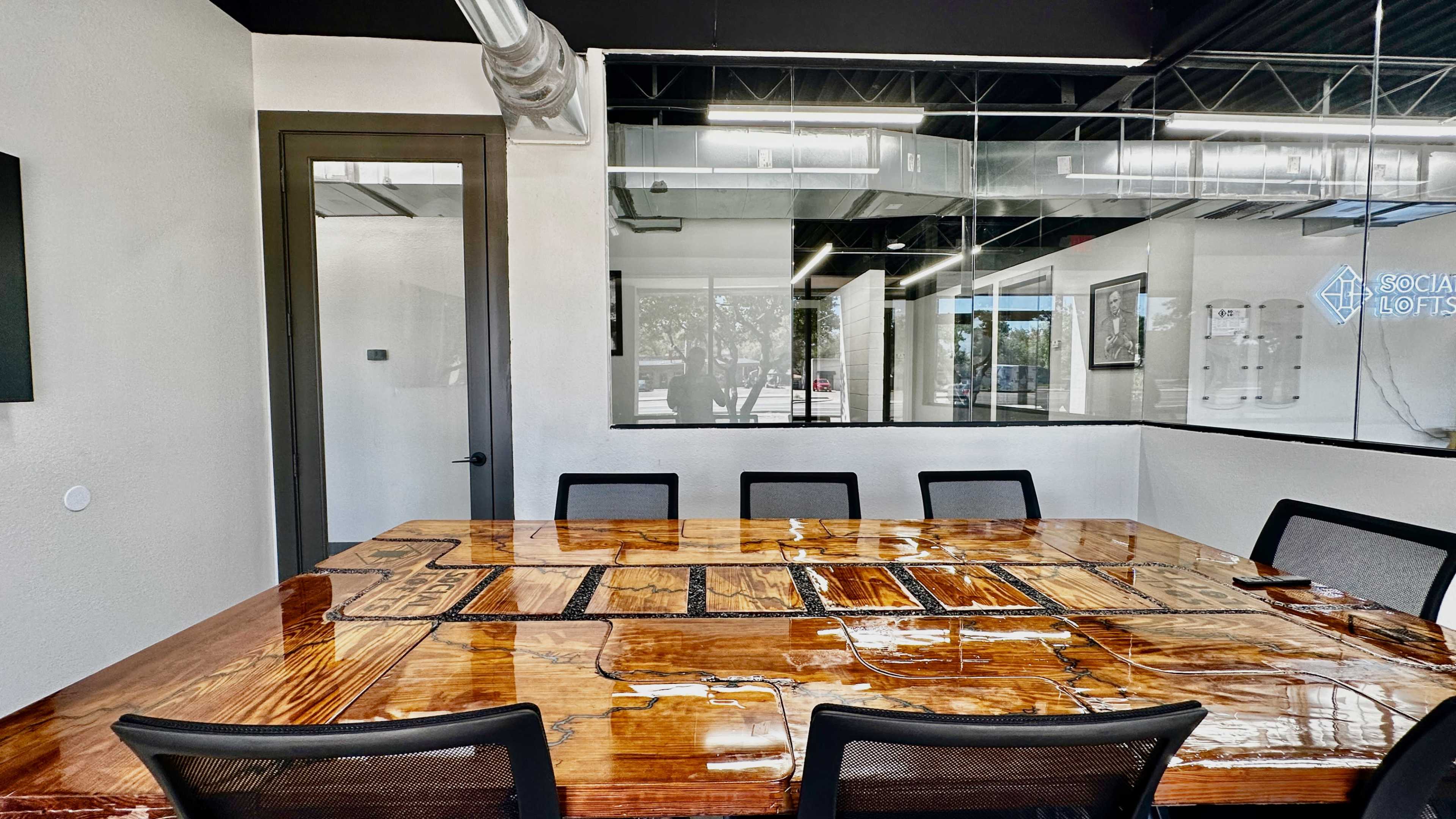The image shows a conference room featuring a large wooden table surrounded by eight black chairs, with glass walls that provide a view of an adjoining space.