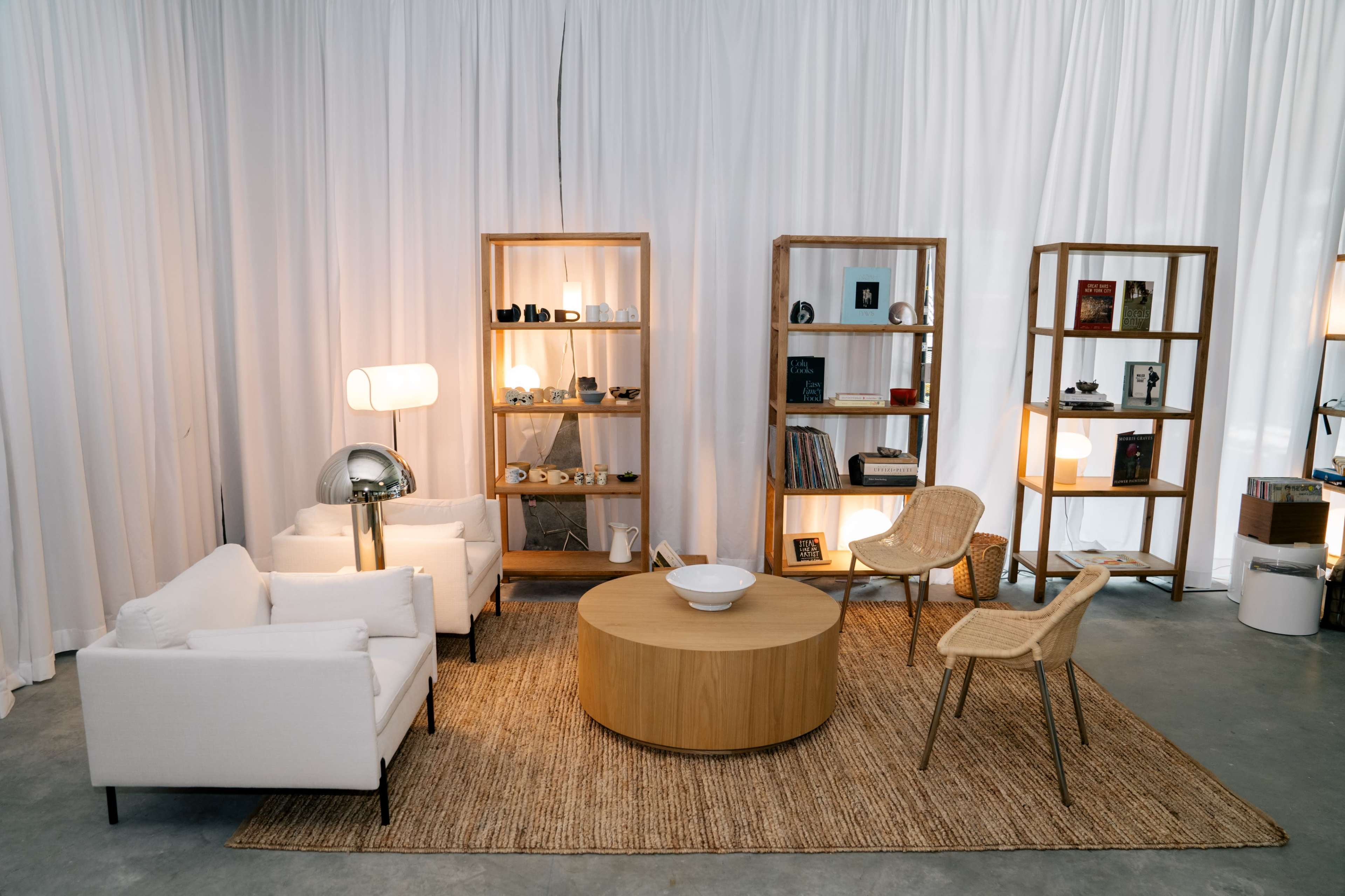 The image shows a modern, minimalistic living room with white curtains, a round wooden coffee table, two white sofas, and three wooden bookshelves displaying various decor items.