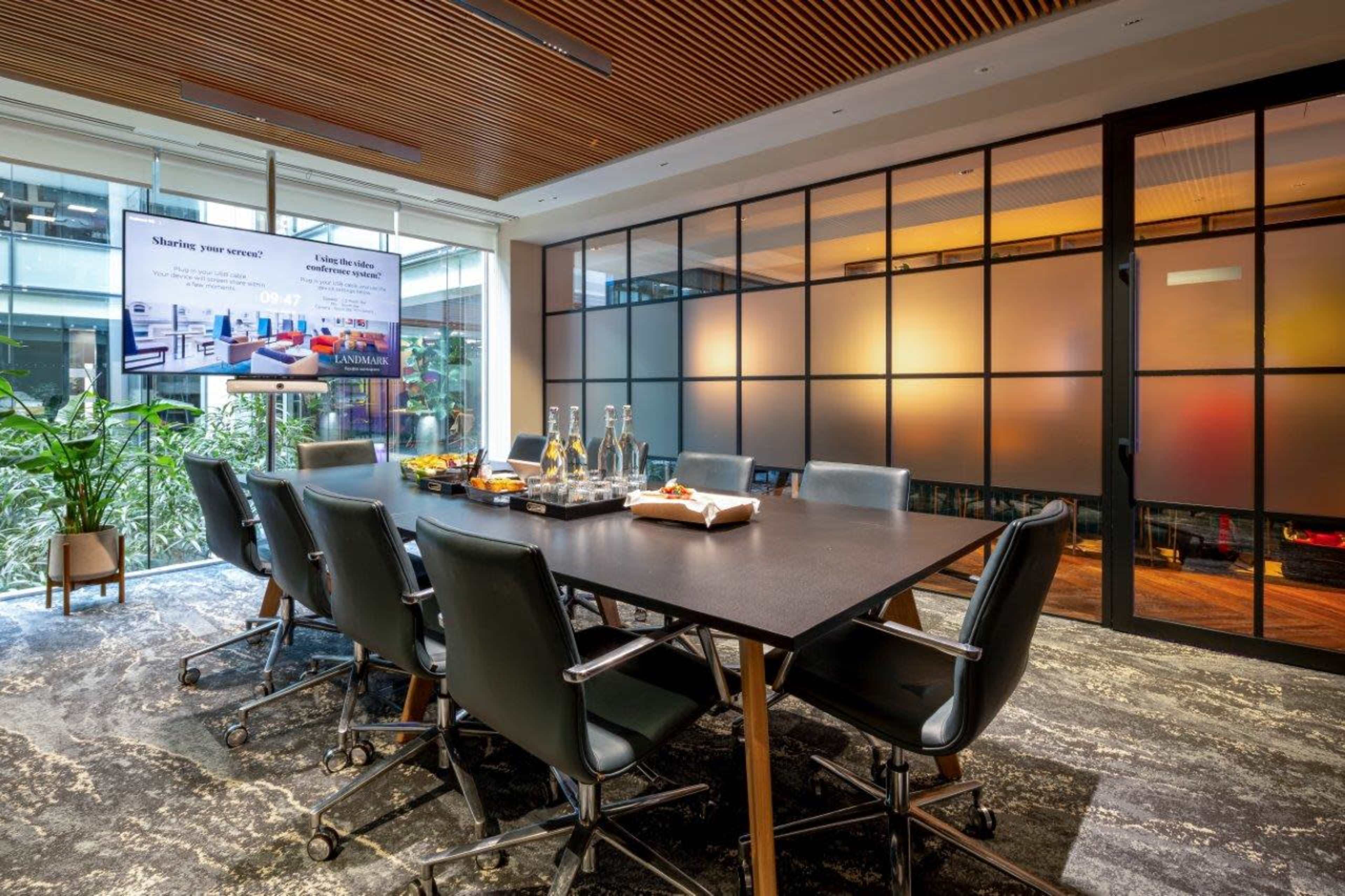 A modern conference room with a large table surrounded by office chairs, a screen displaying a presentation, and glass walls providing a view of an indoor plant and natural light.