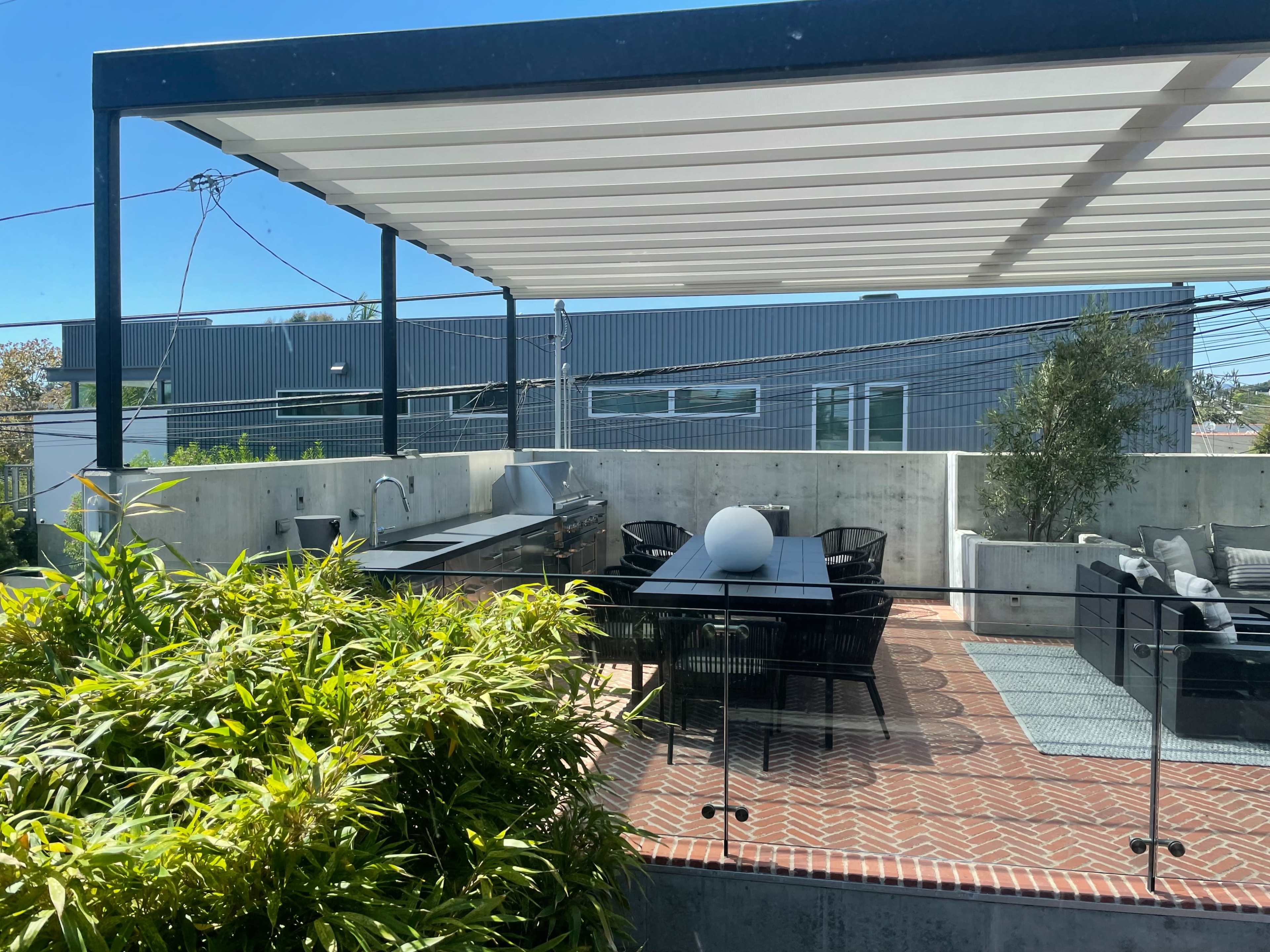 A modern outdoor patio with a dining area, a grill, and planters, all under a pergola with a shaded roof.
