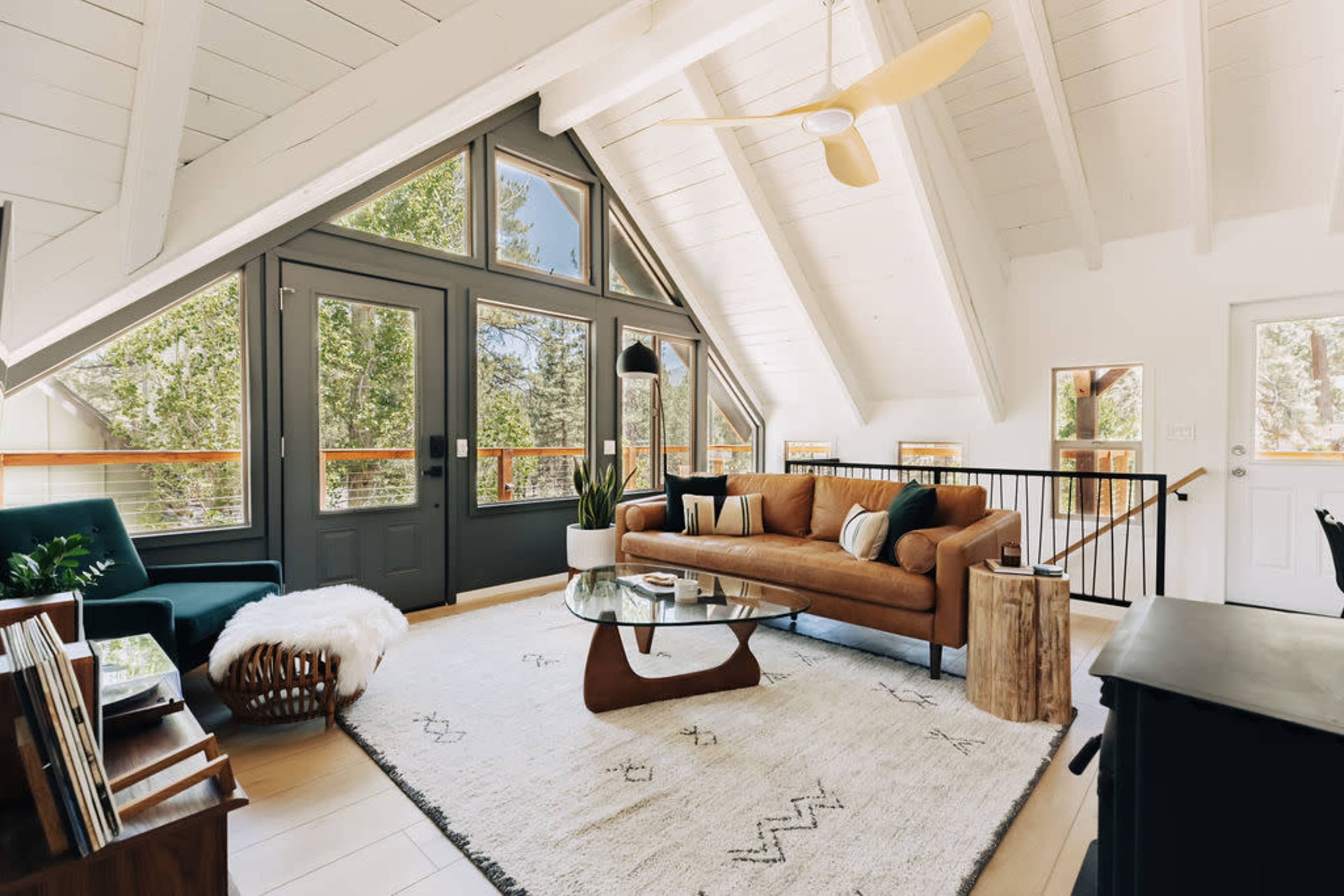 A modern living room features a brown sofa, a glass coffee table, and large windows framed in dark blue, set under a vaulted ceiling with wooden beams.