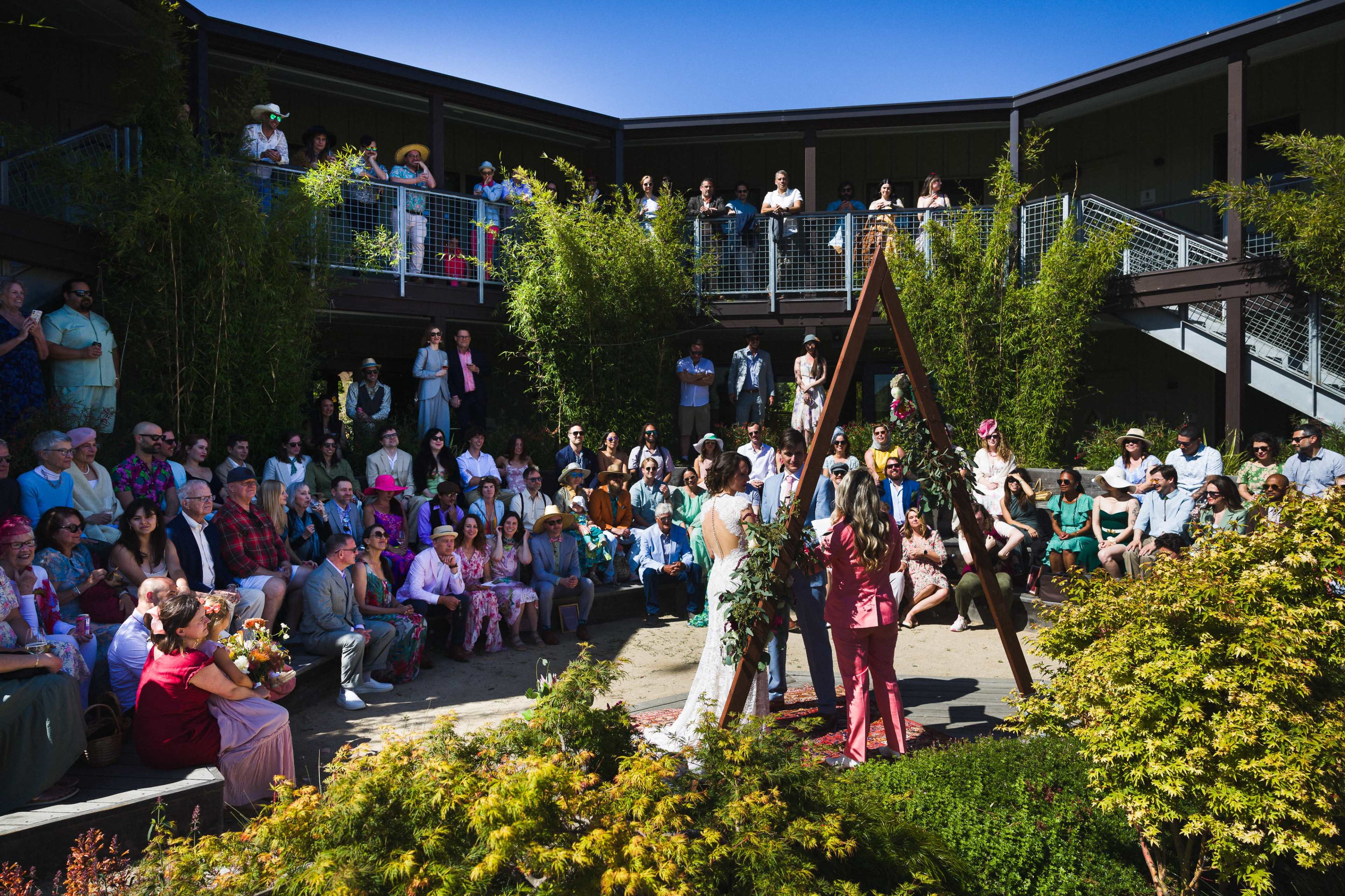 A wedding ceremony is taking place outdoors, with a diverse group of guests seated around the couple standing beneath an arch.
