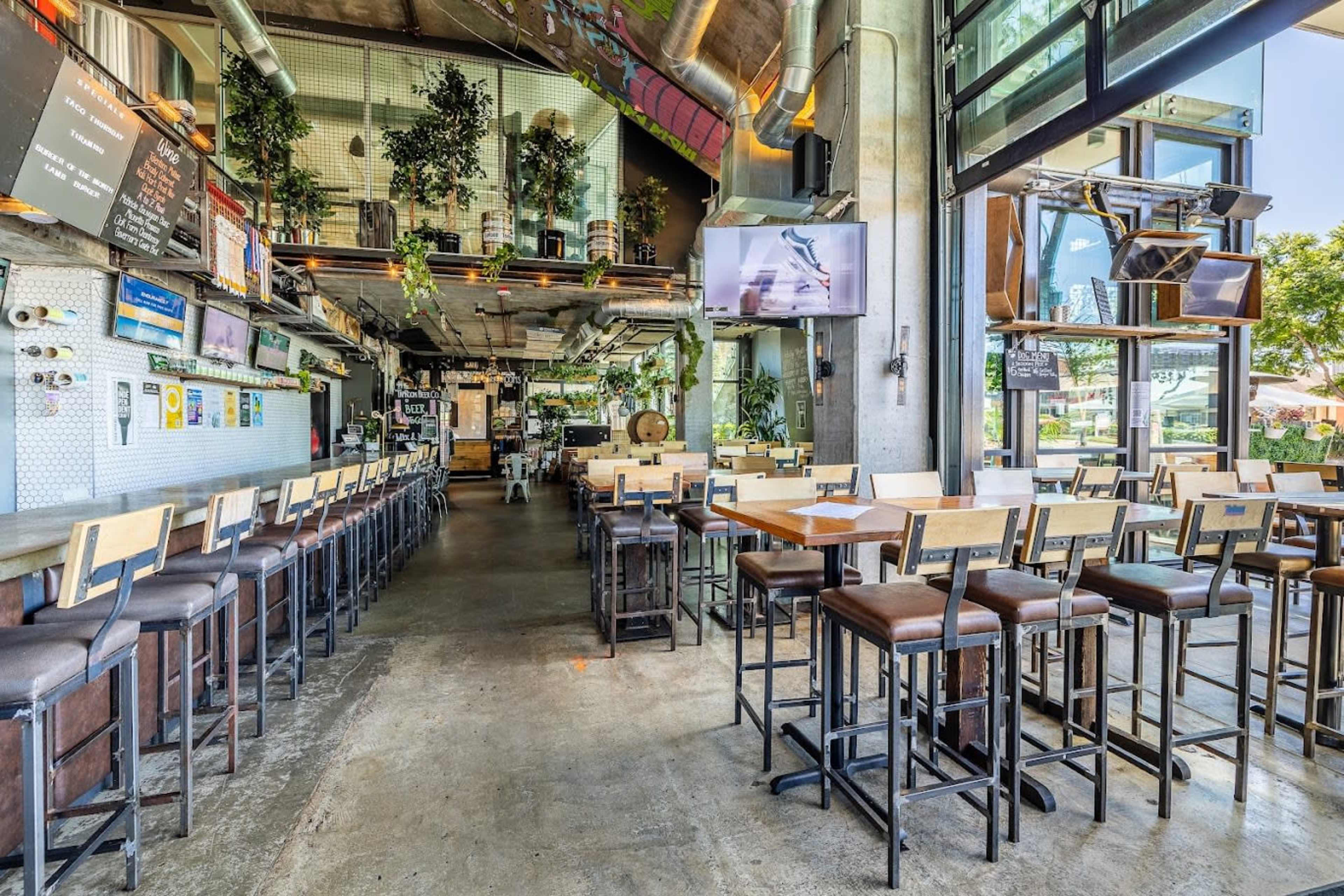 A spacious, modern restaurant interior with tall bar stools along a counter and a row of tables arranged for dining.
