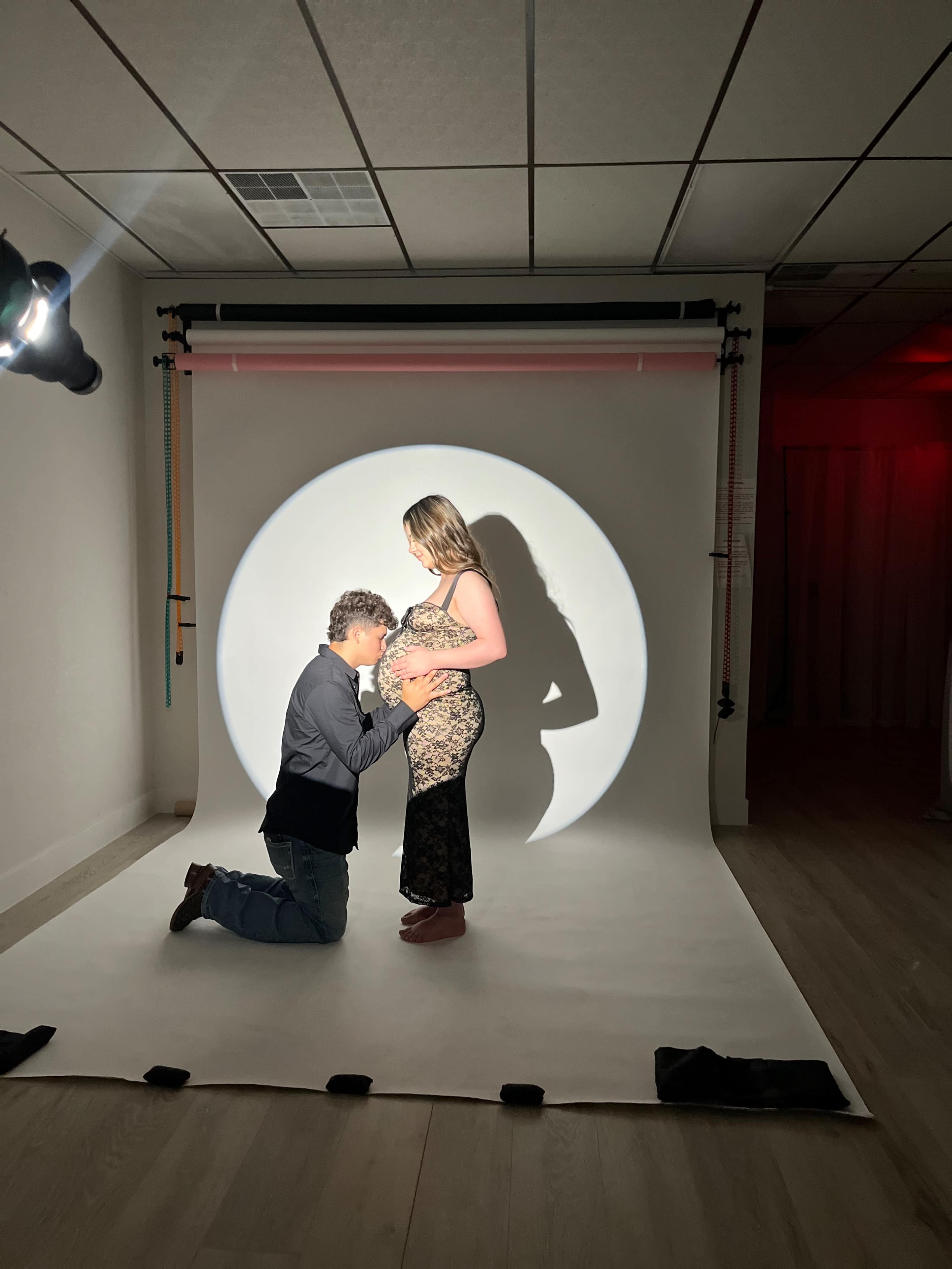 A man kneels in front of a pregnant woman while they are both illuminated by a circular light on a backdrop in a studio.