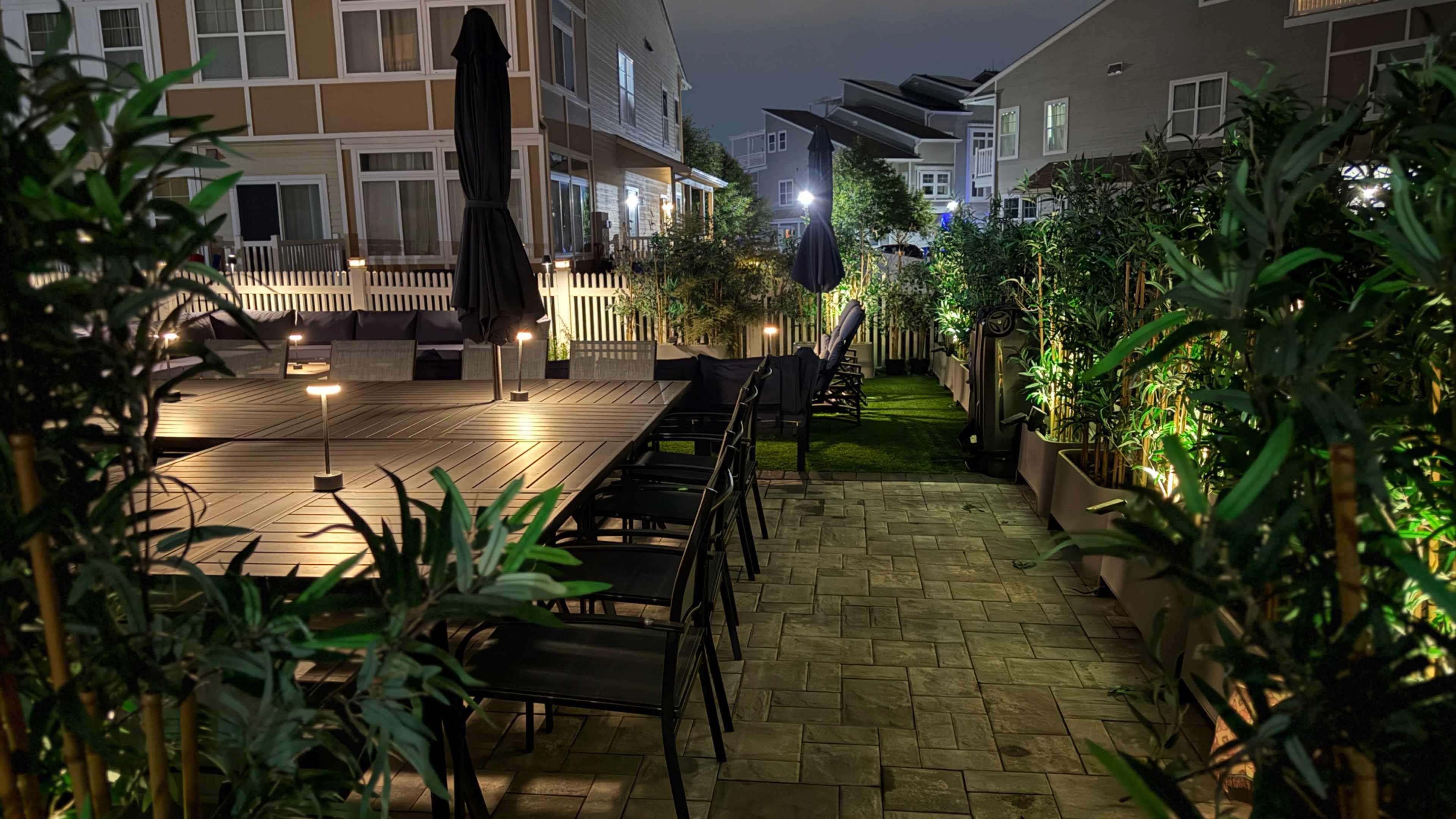 The image shows a dimly lit outdoor patio area with a large table, chairs, and decorative plants lining either side of a pathway leading to more seating and lanterns.