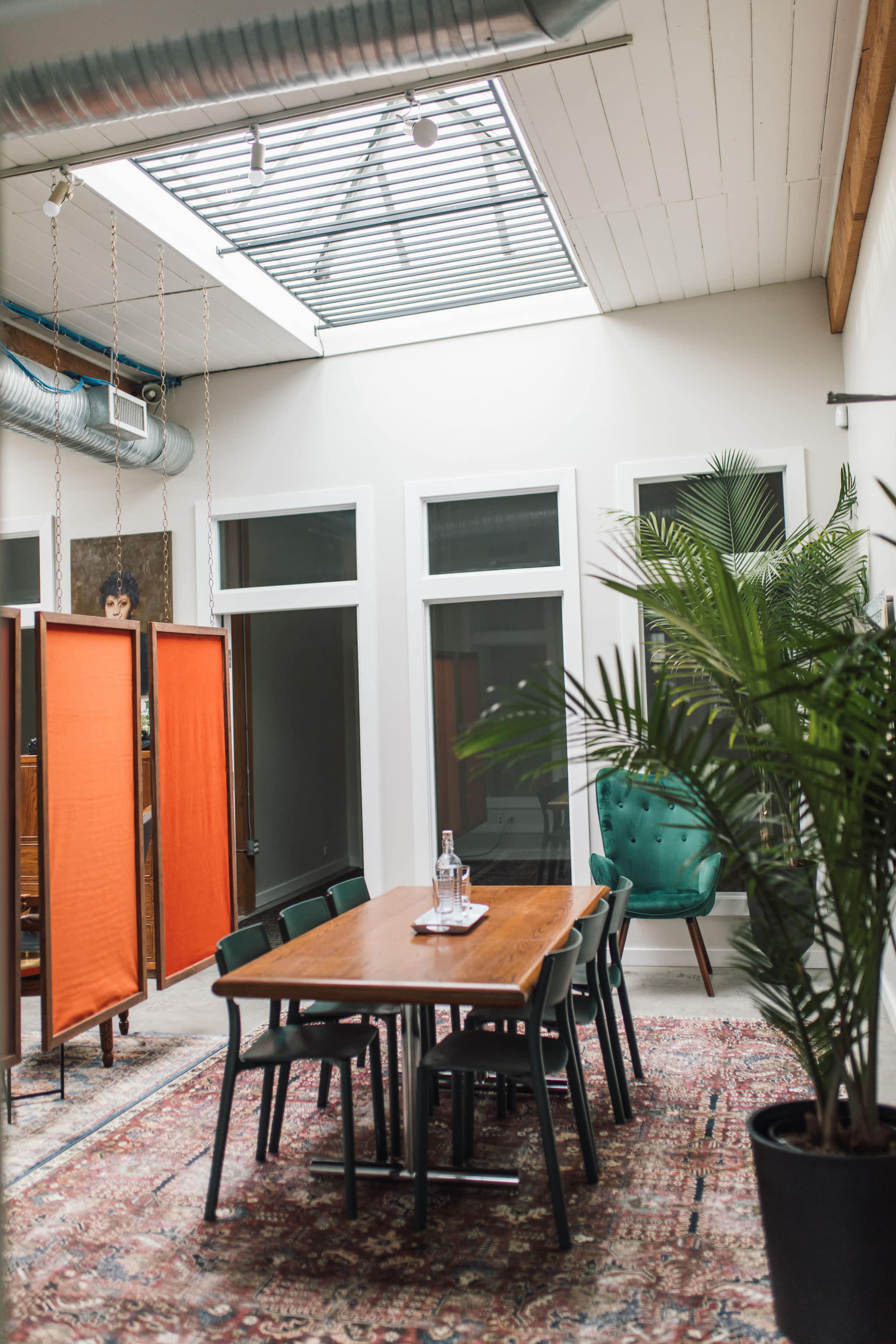 The image shows a modern dining area with a wooden table surrounded by chairs, a large indoor plant, and bright natural light coming through a skylight.