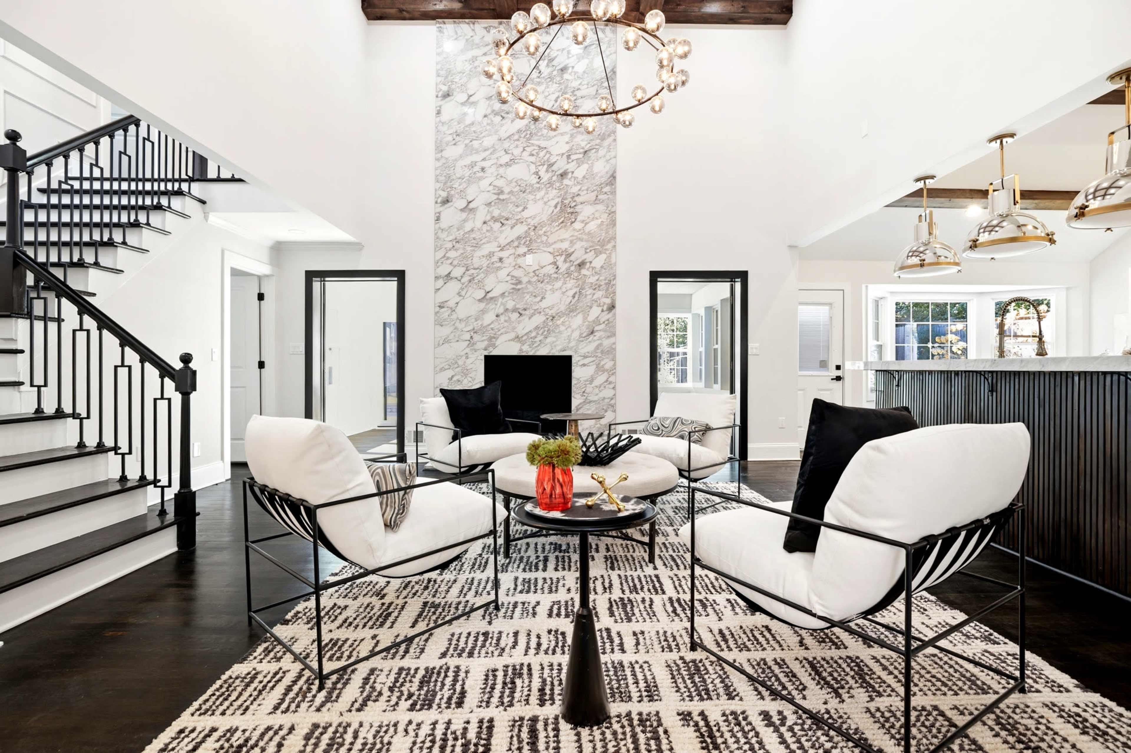 The image shows a modern living room with a marble feature wall, a black metal table surrounded by upholstered chairs, and a staircase in the background.