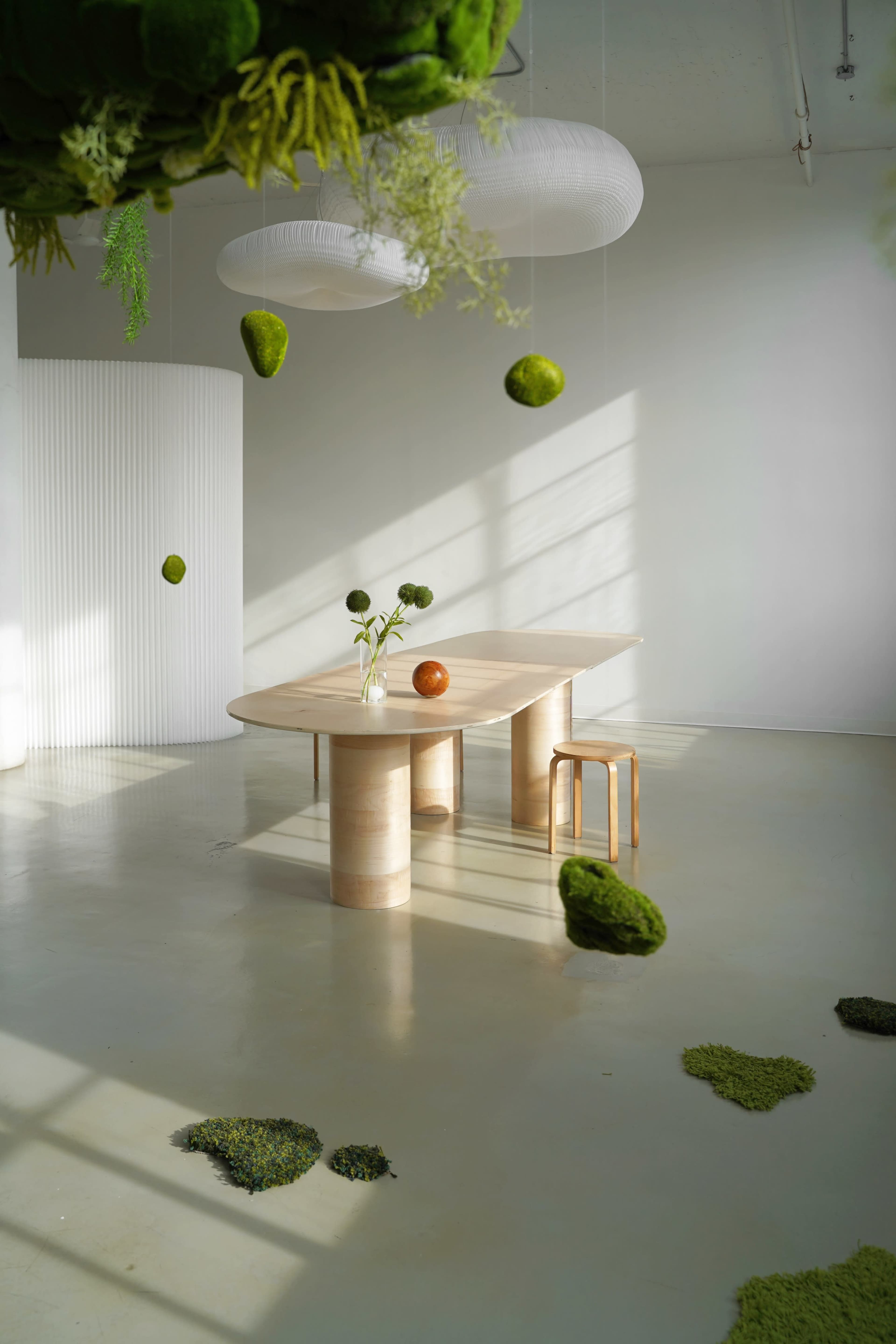 A minimalist interior features a large wooden table with a single stool, surrounded by suspended moss and a few decorative elements on the floor.