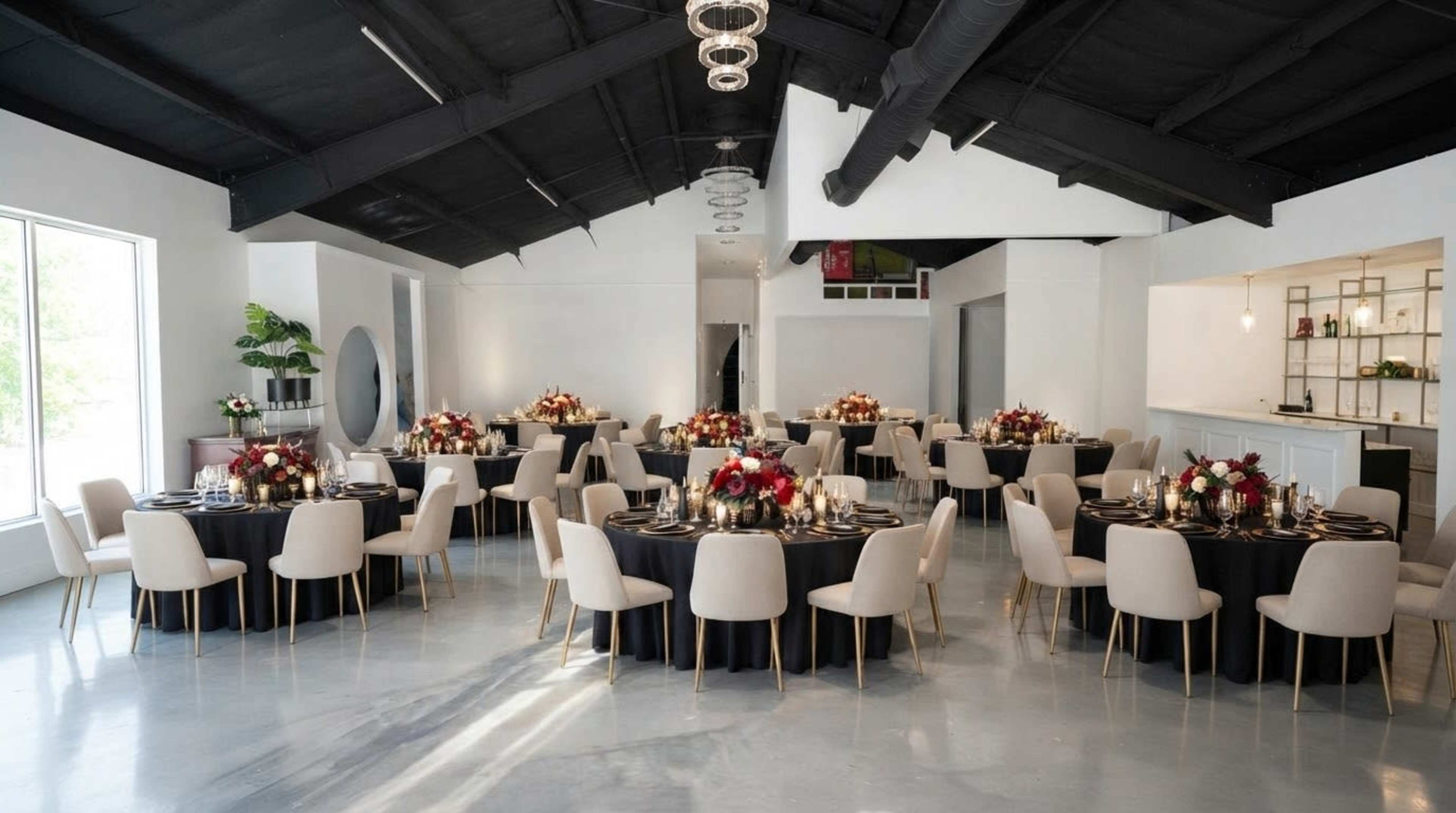 A modern event space features round tables set with floral centerpieces and elegant dinnerware, surrounded by neutral-colored chairs under a high black ceiling.