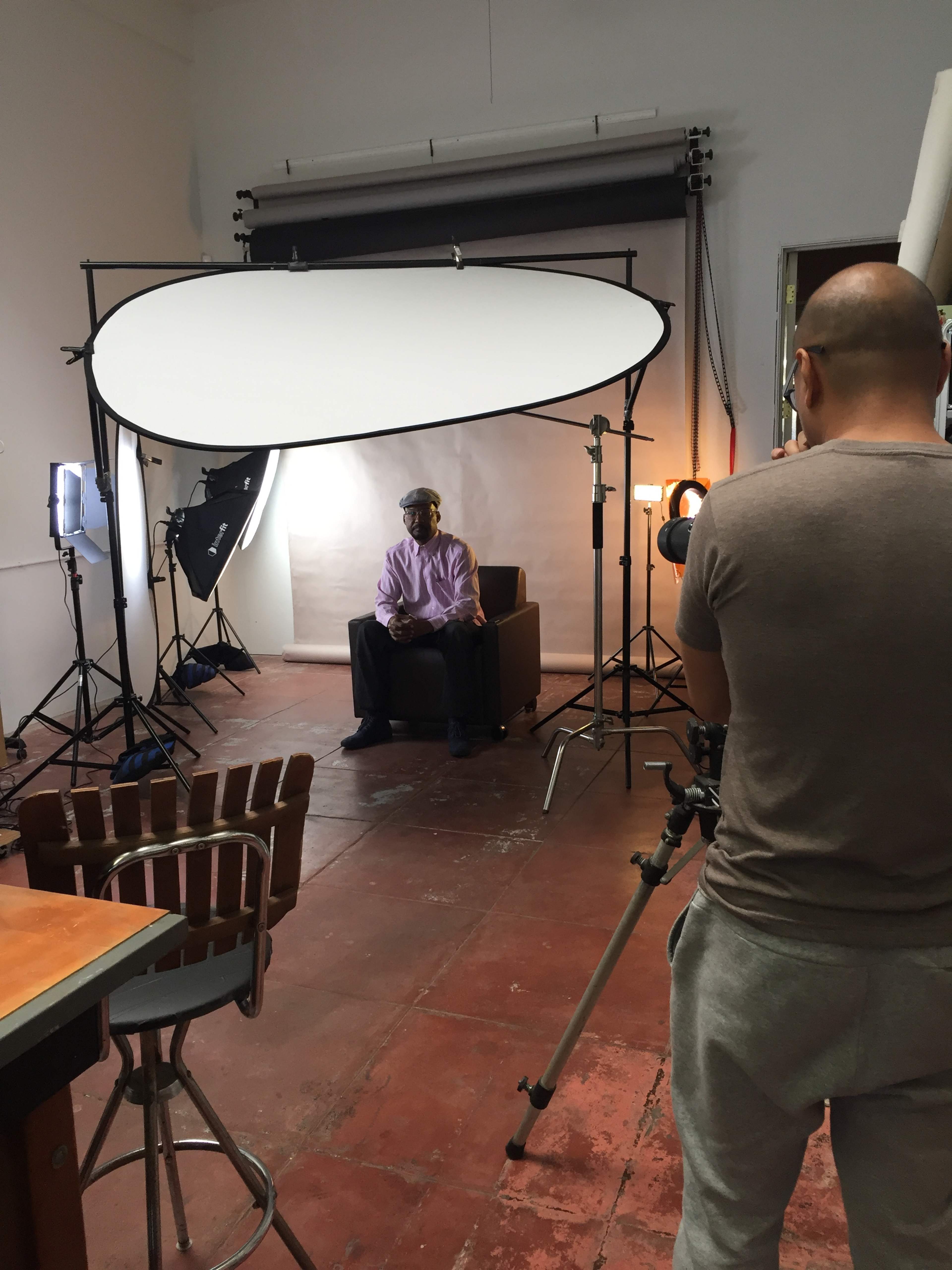 A man wearing a cap sits on a couch in a studio setting, while another person operates a camera to the right.