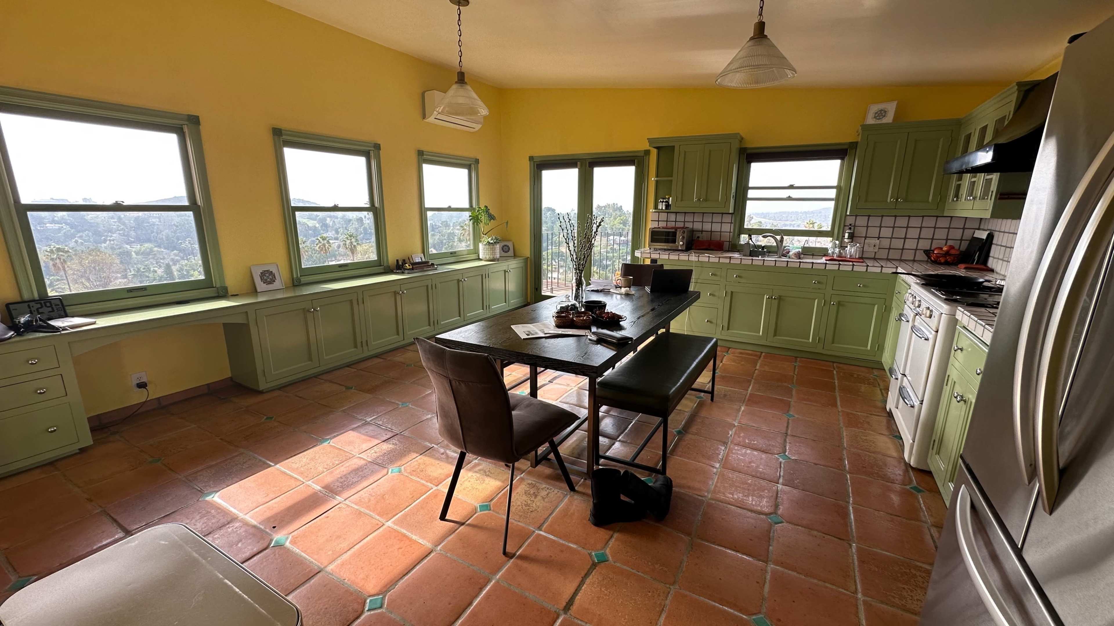 The kitchen features green cabinetry, a large black dining table, and terracotta tile flooring, with windows providing a view of the surrounding landscape.