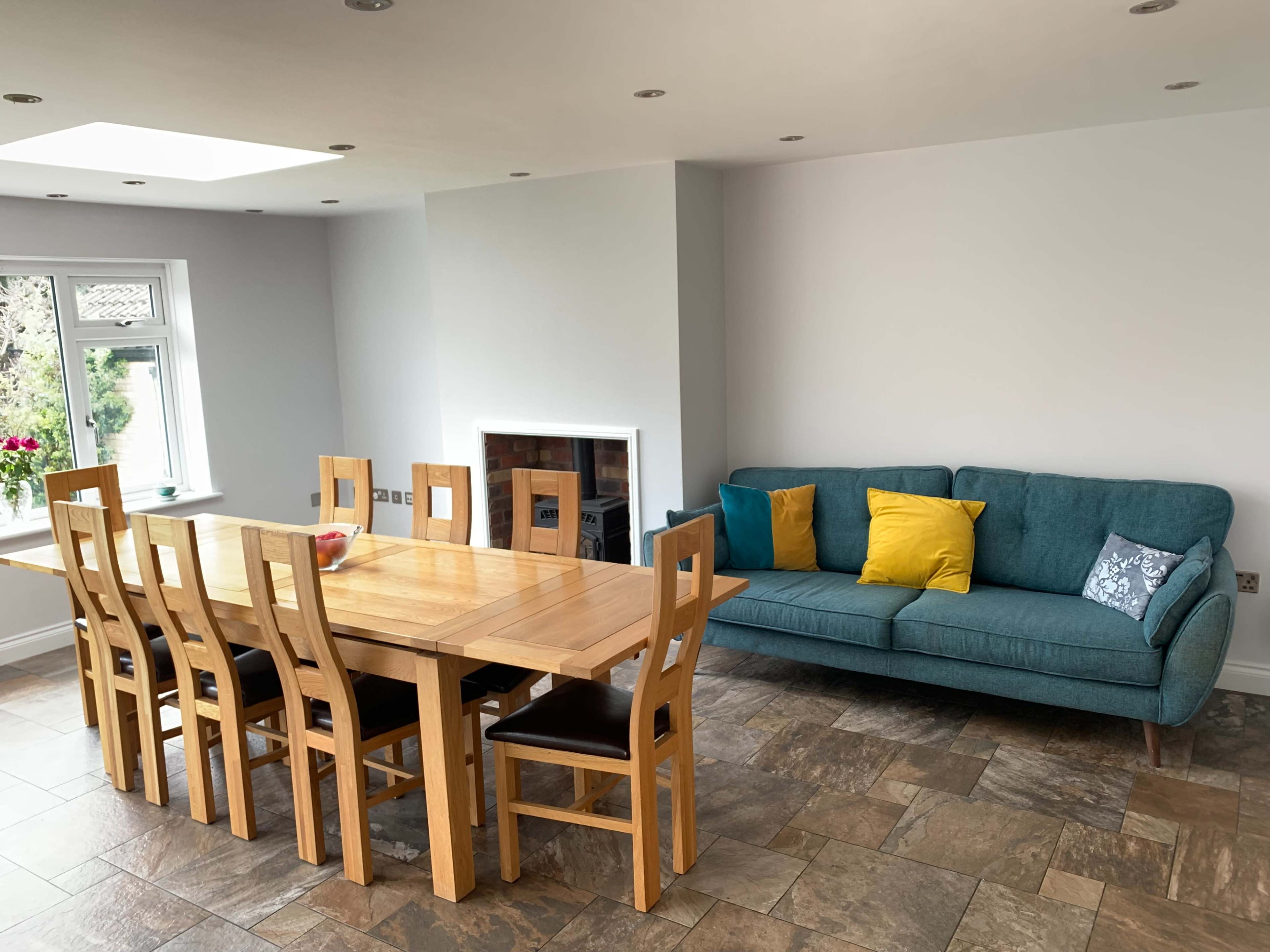 The image shows a modern dining area featuring a large wooden table with eight chairs, a blue sofa with colorful cushions, and a window that allows natural light to enter the space.