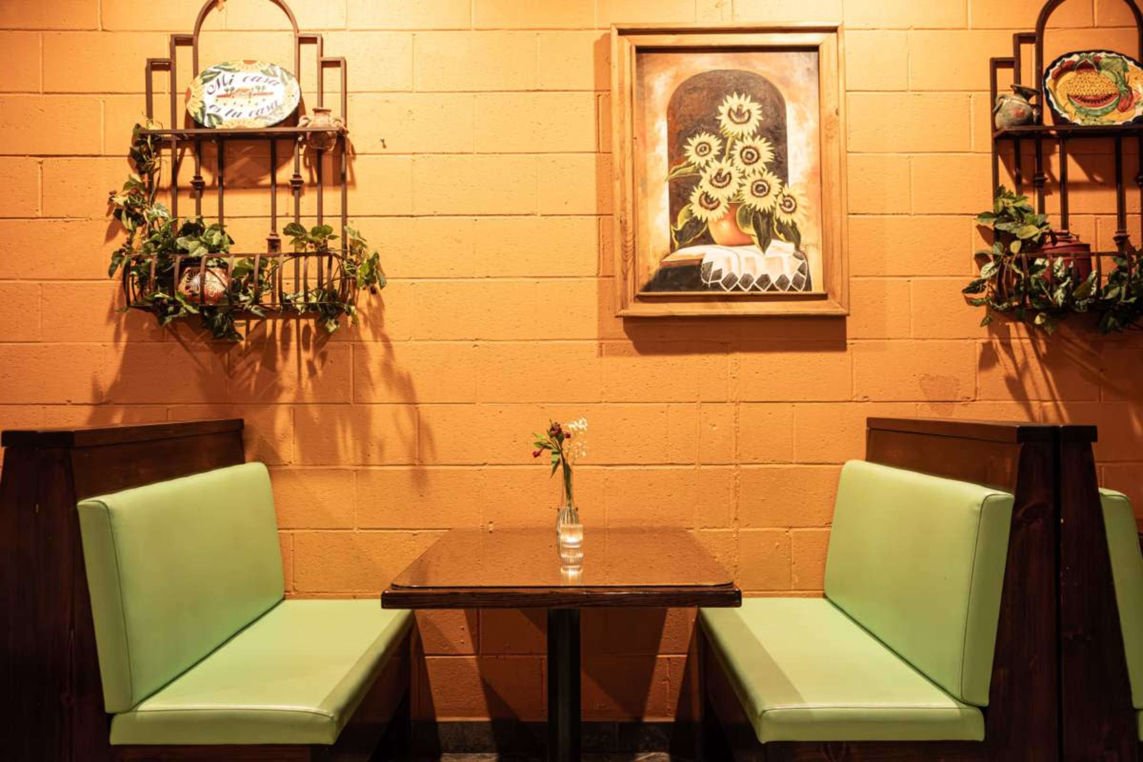 A small table with a single flower vase sits between two green upholstered benches in a cozy restaurant, adorned with a wall painting and decorative shelves.